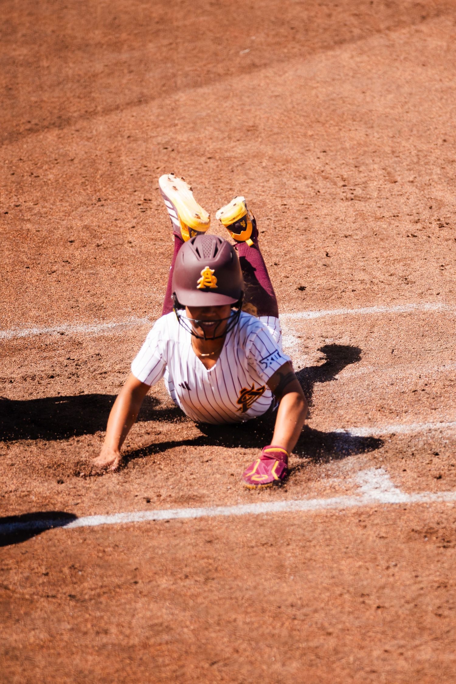 Baseball player in maroon uniform sliding into base on dirt infield during a sunny game.