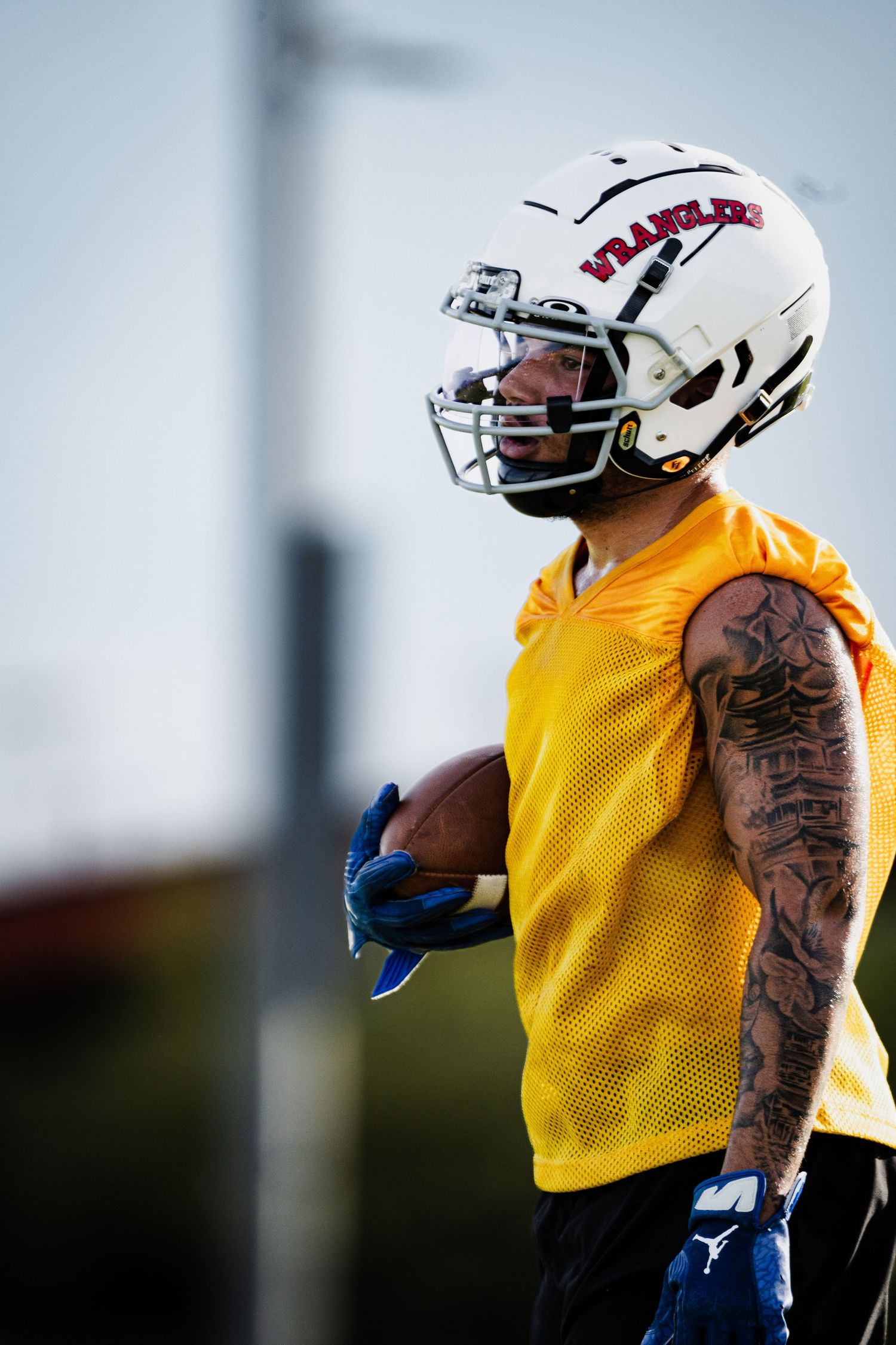 A tattooed football player in a yellow jersey and white Wranglers helmet holds a football during practice.