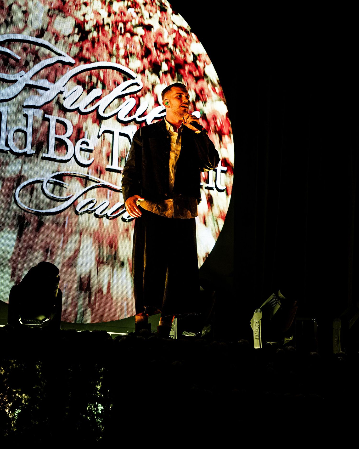 A performer stands on a dark stage with a large illuminated backdrop displaying tour text during a live concert.