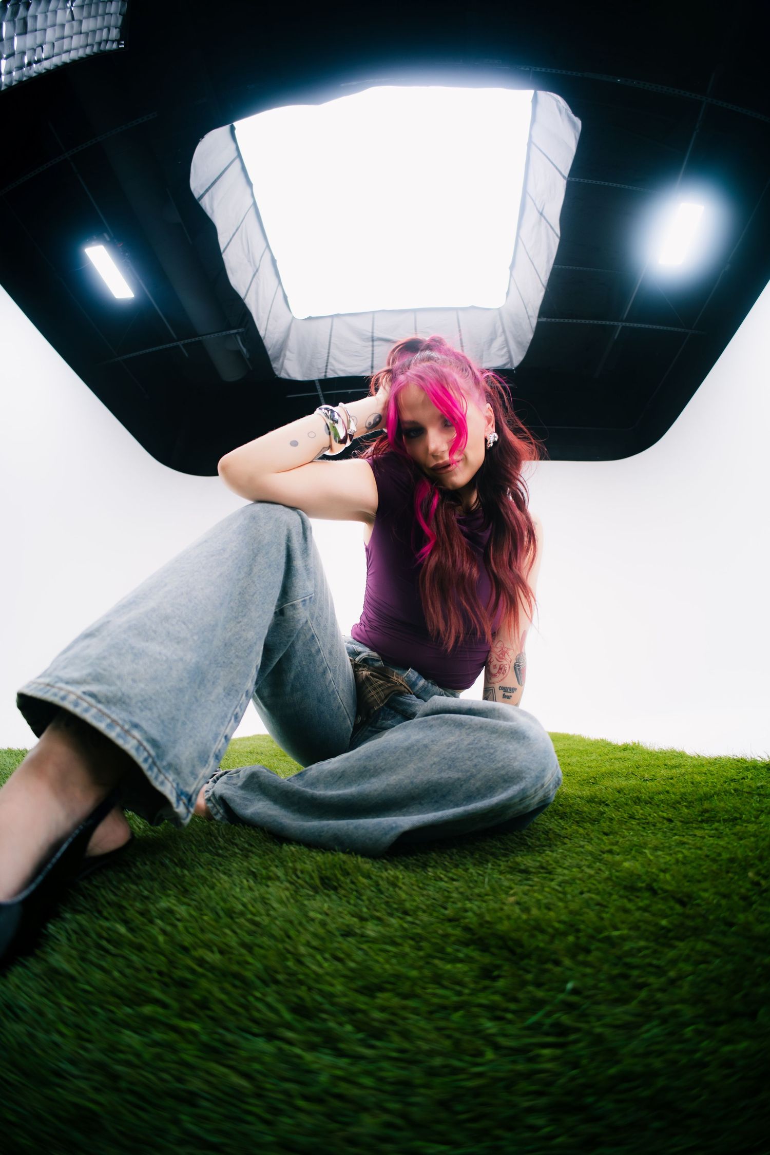 Young woman with pink hair sitting on grass in a photography studio, wearing wide-leg jeans and a purple top.
