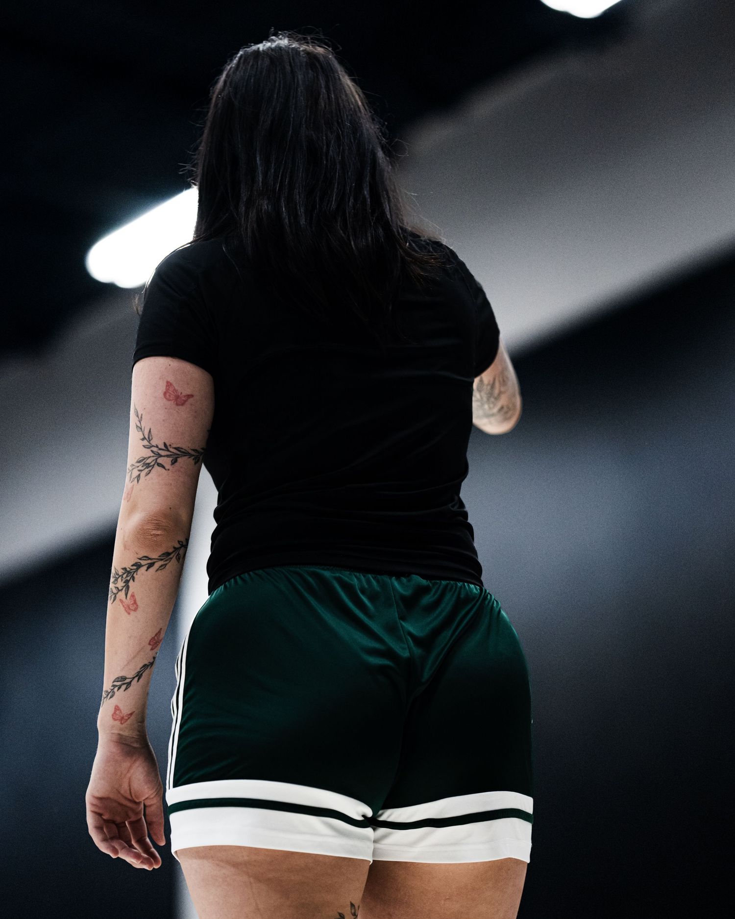 A woman with tattoos wearing green shorts and a black top stands on a basketball court.