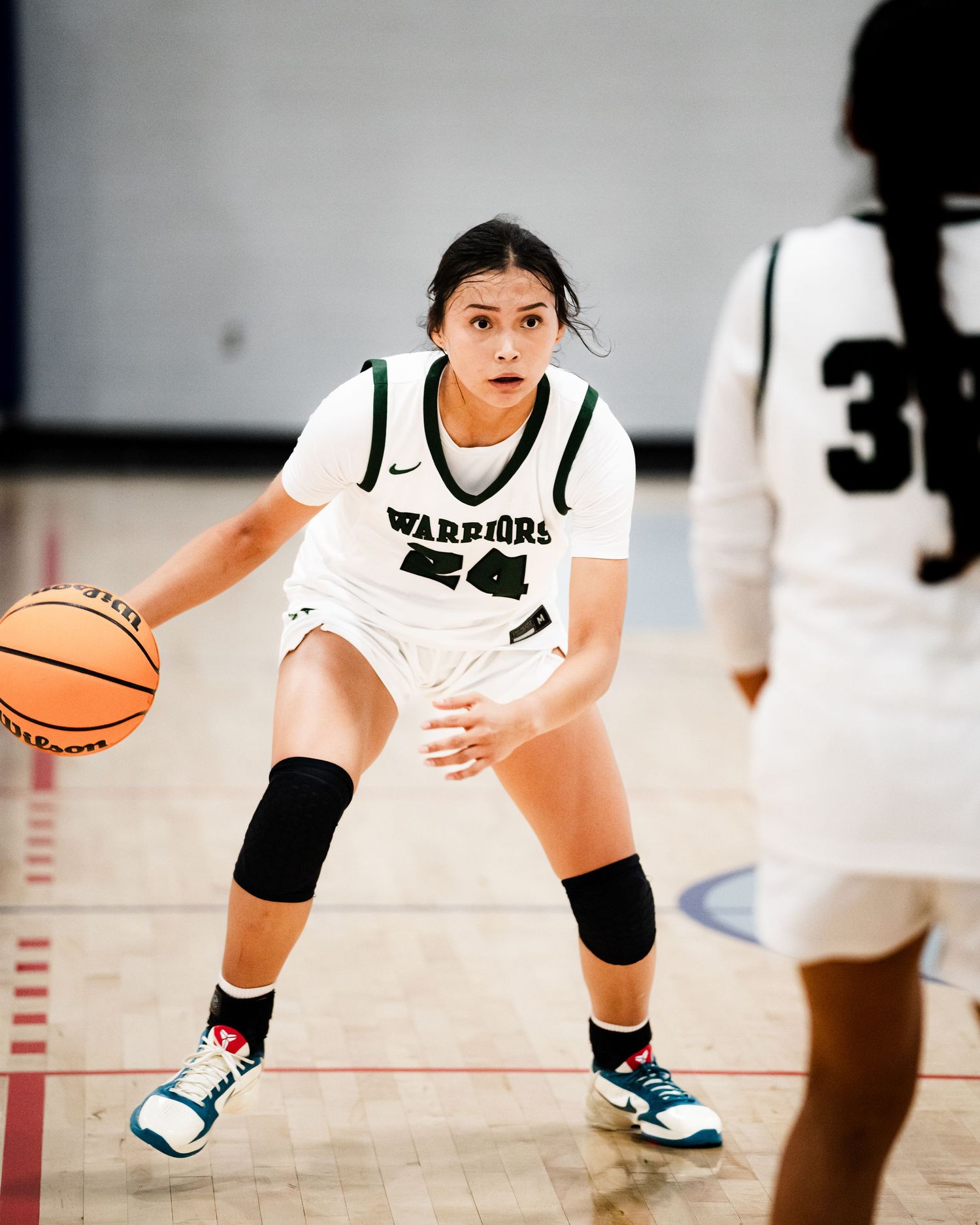 A female Warriors number 24 basketball player in white dribbles the ball while defending on the court.