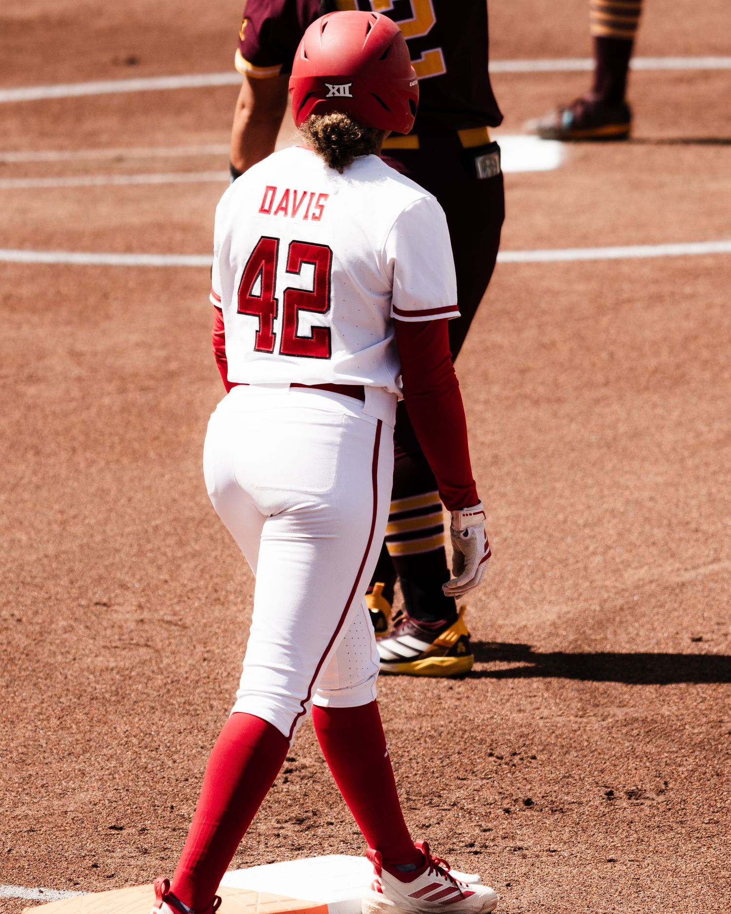 Softball player Davis #42 in white and red uniform stands on base during a sunny game.