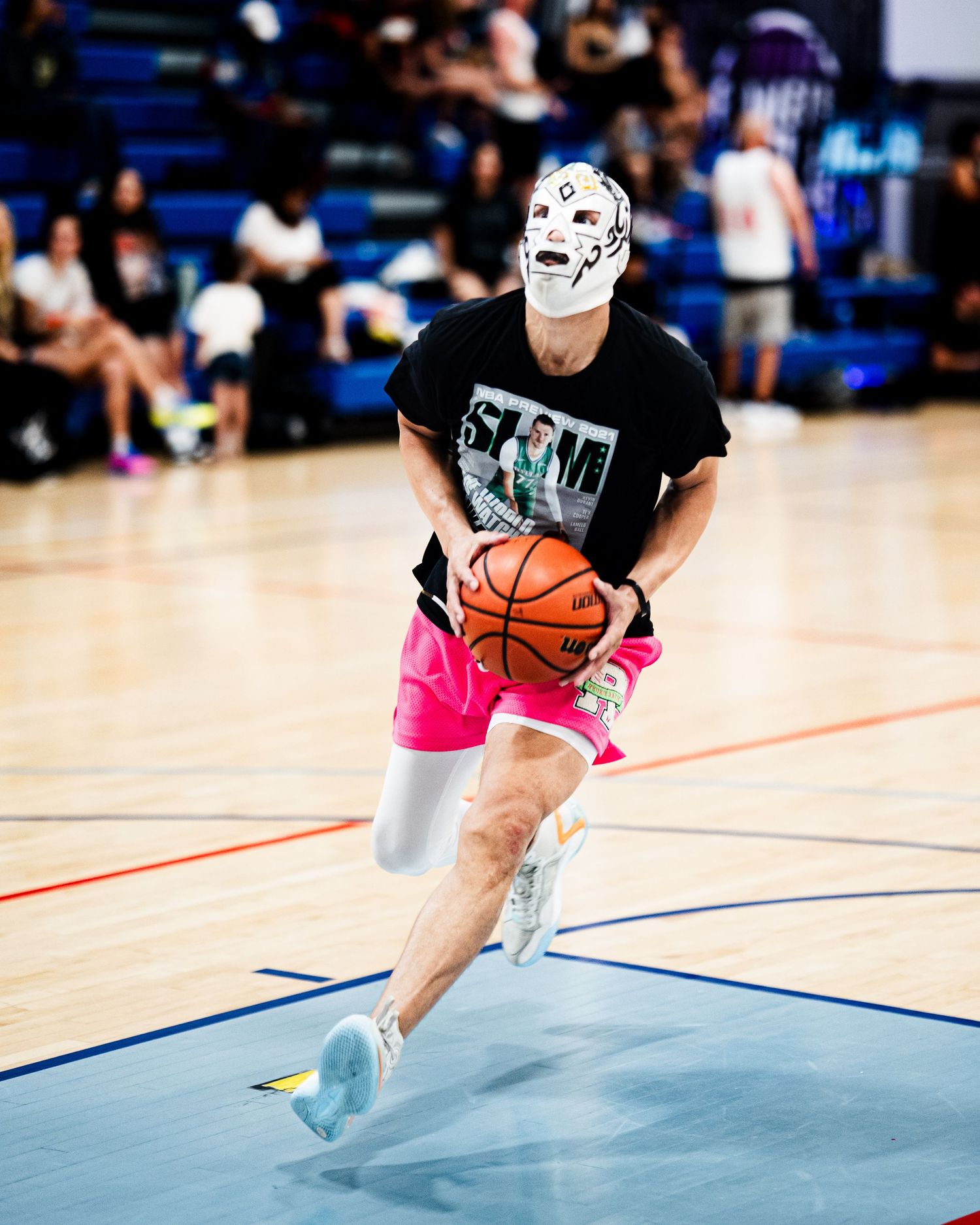 Player wearing a white mask and pink shorts dribbles a basketball on an indoor court during a game.