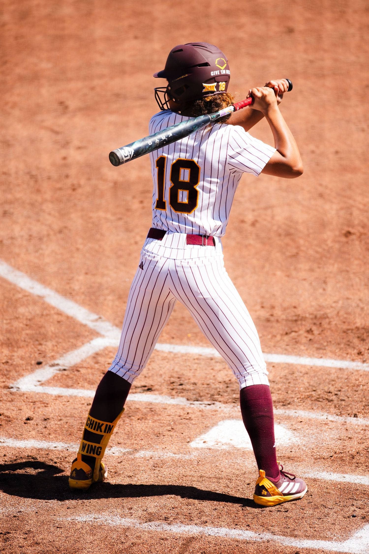 Softball player #18 in white pinstripe uniform stands in batting stance on dirt field, bat raised and ready to swing.