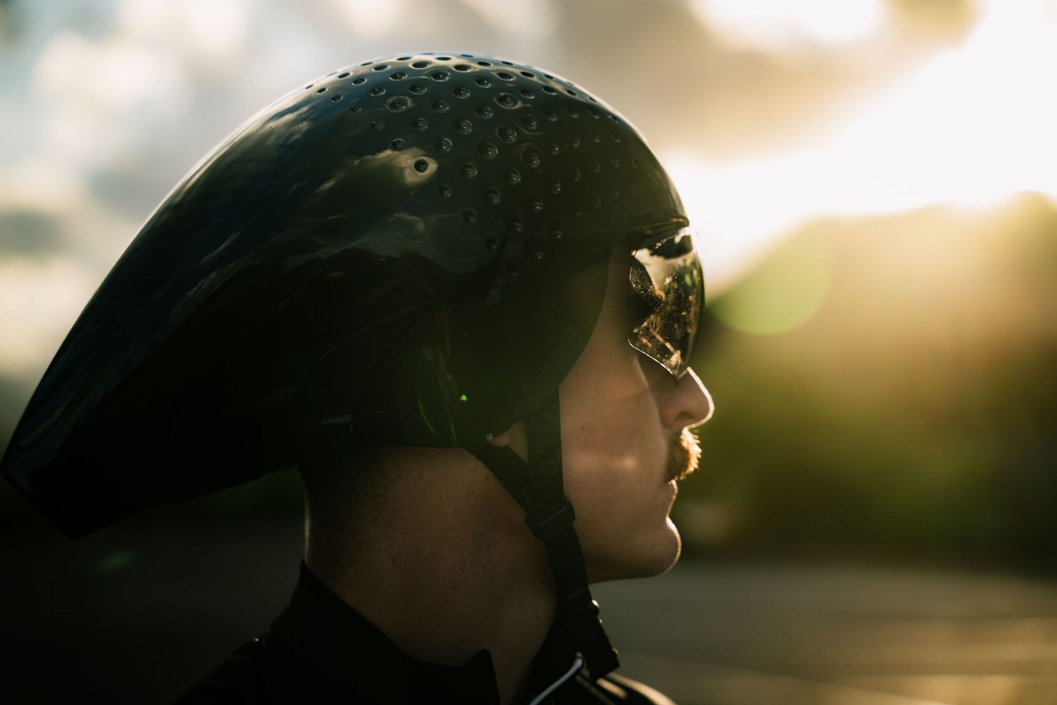 Person wearing a black motorcycle helmet and goggles with golden sunset light reflecting off the visor outdoors.