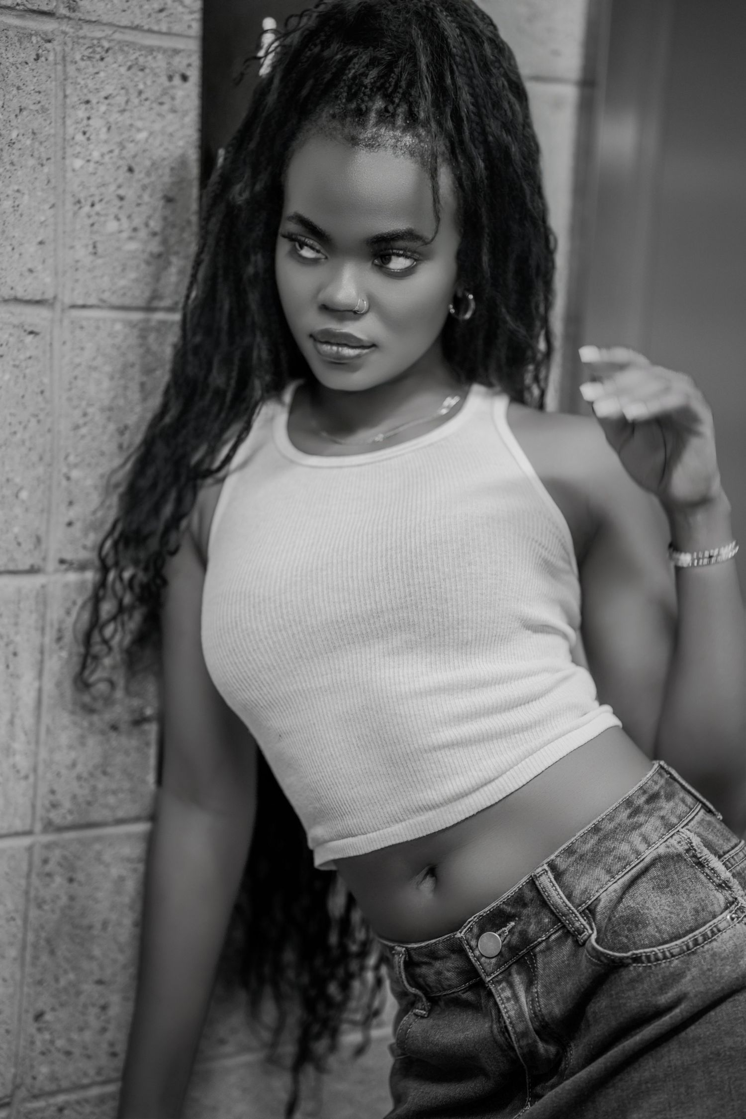 Black and white portrait of a young woman with long braids wearing a crop top and jeans leaning against a brick wall.