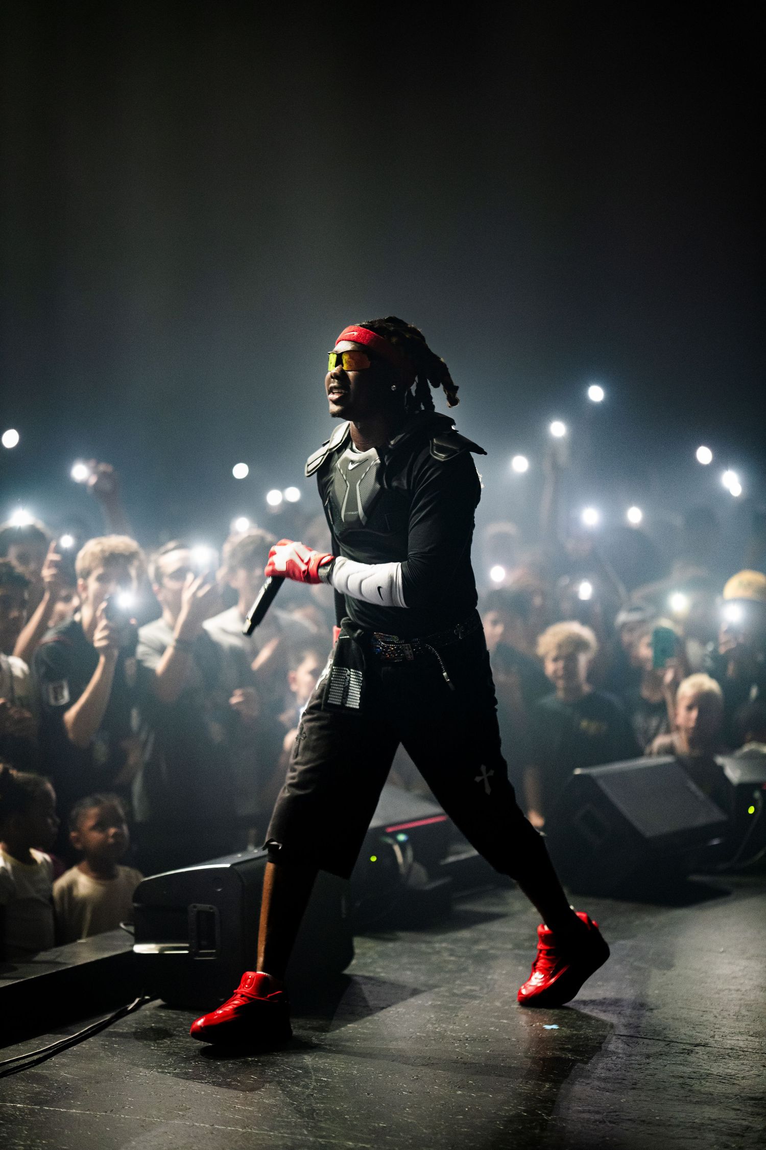 Rapper performing on stage in red sneakers and headband, holding microphone with crowd lights glowing behind.