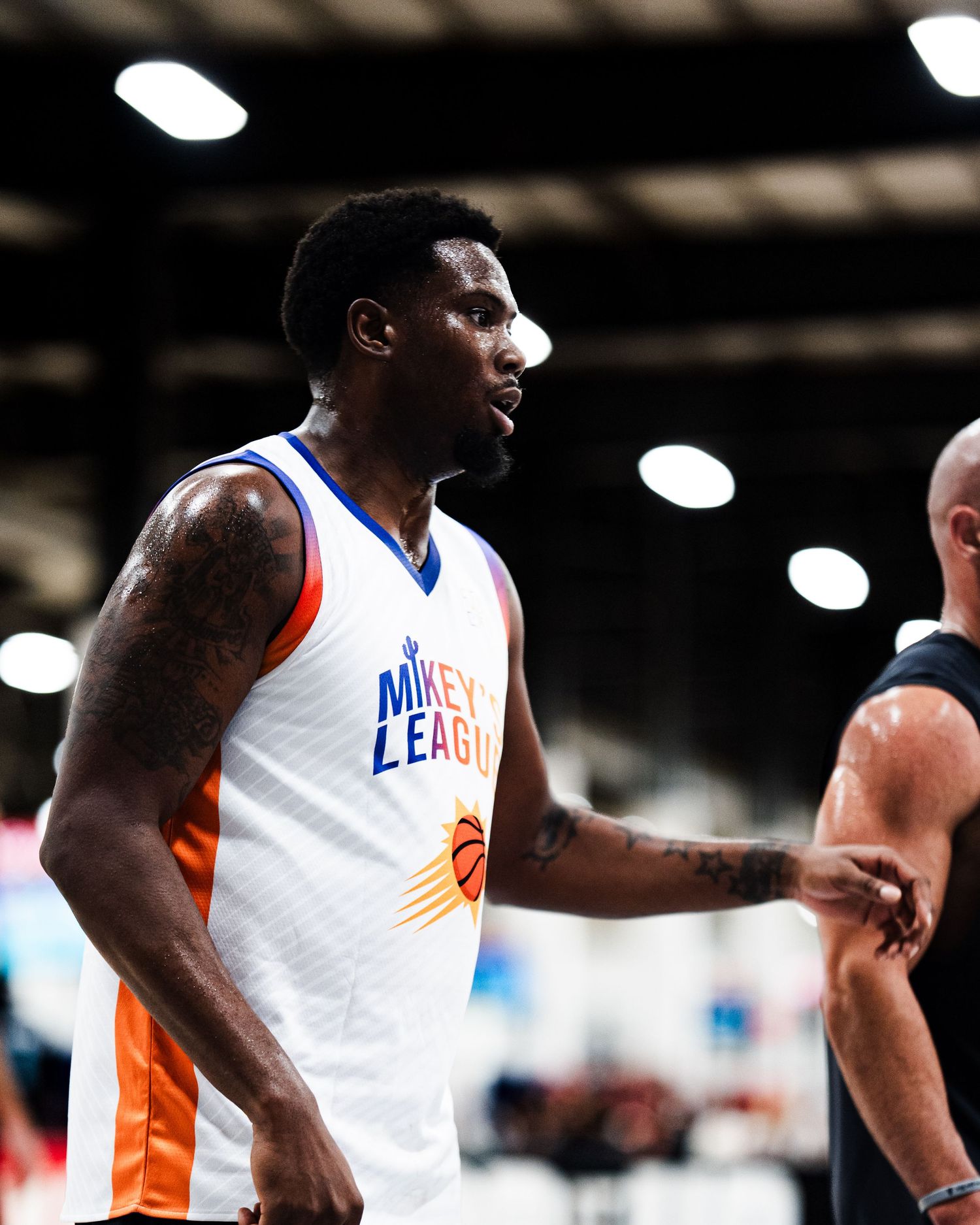 A tattooed basketball player in a white Mikeys League jersey stands on an indoor court during a game.