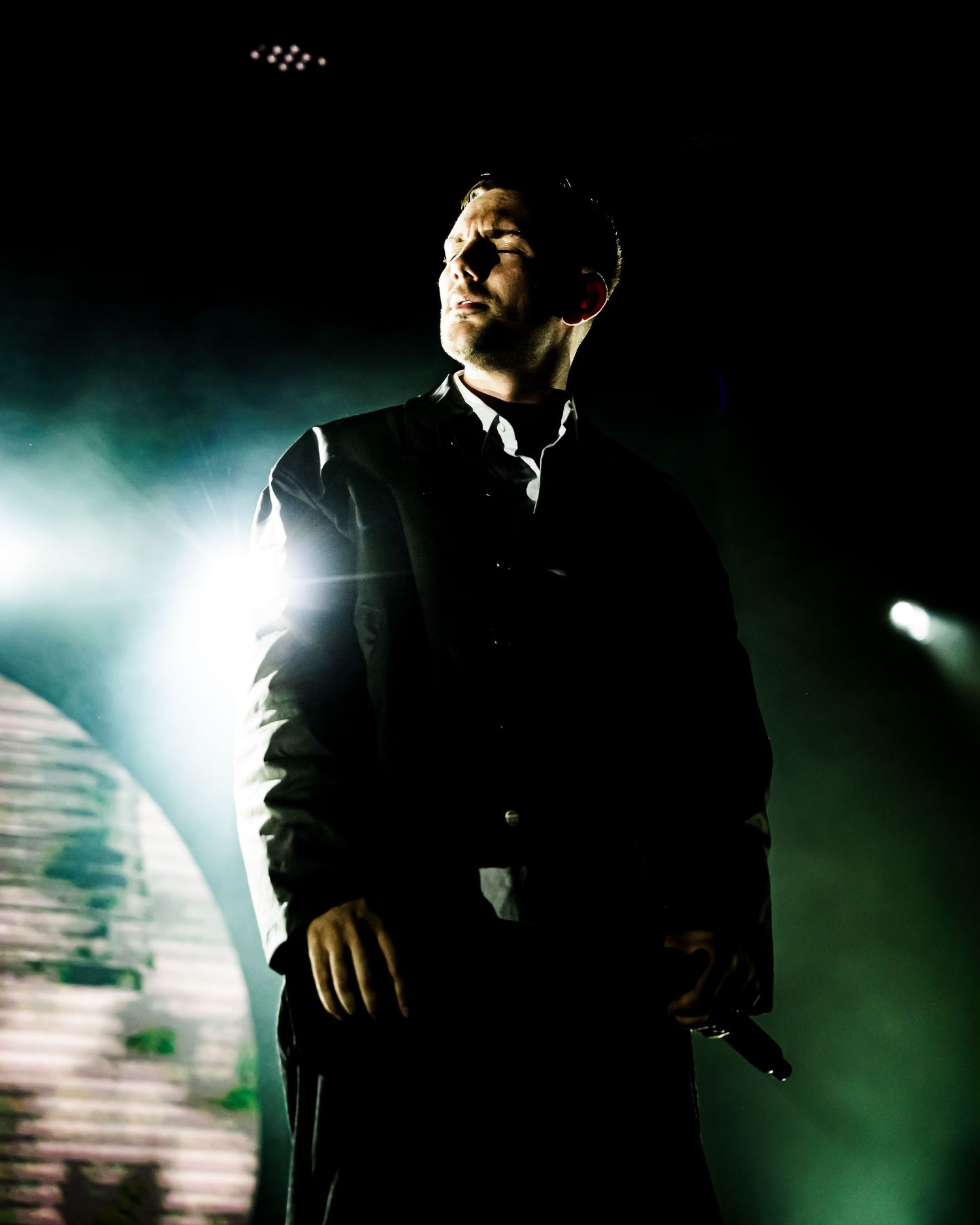 A dramatic backlit male performer on stage in a dark suit, holding a microphone with green stage lights behind him.