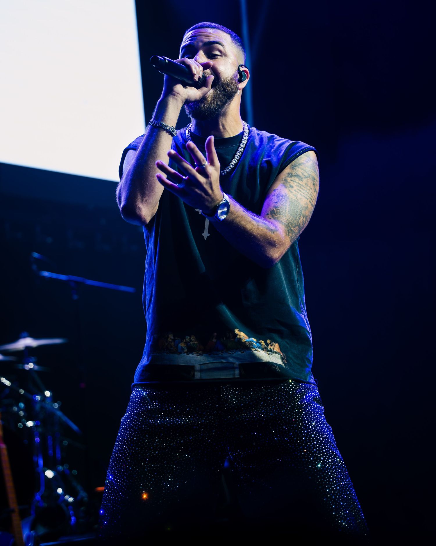 A bearded male performer sings into a microphone on stage wearing a sleeveless shirt and sparkling blue pants.