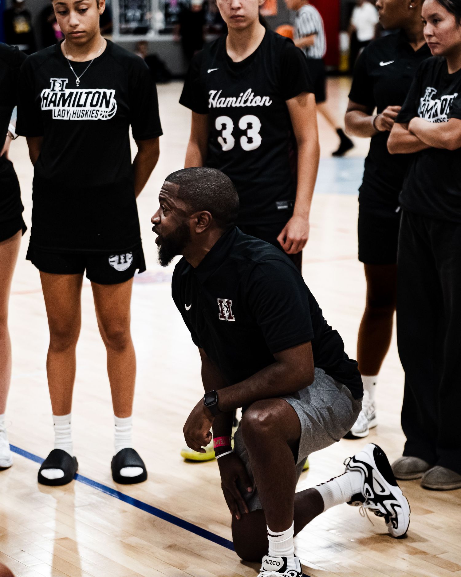 Male basketball coach kneels on court giving instructions to Hamilton Lady Huskies team players.