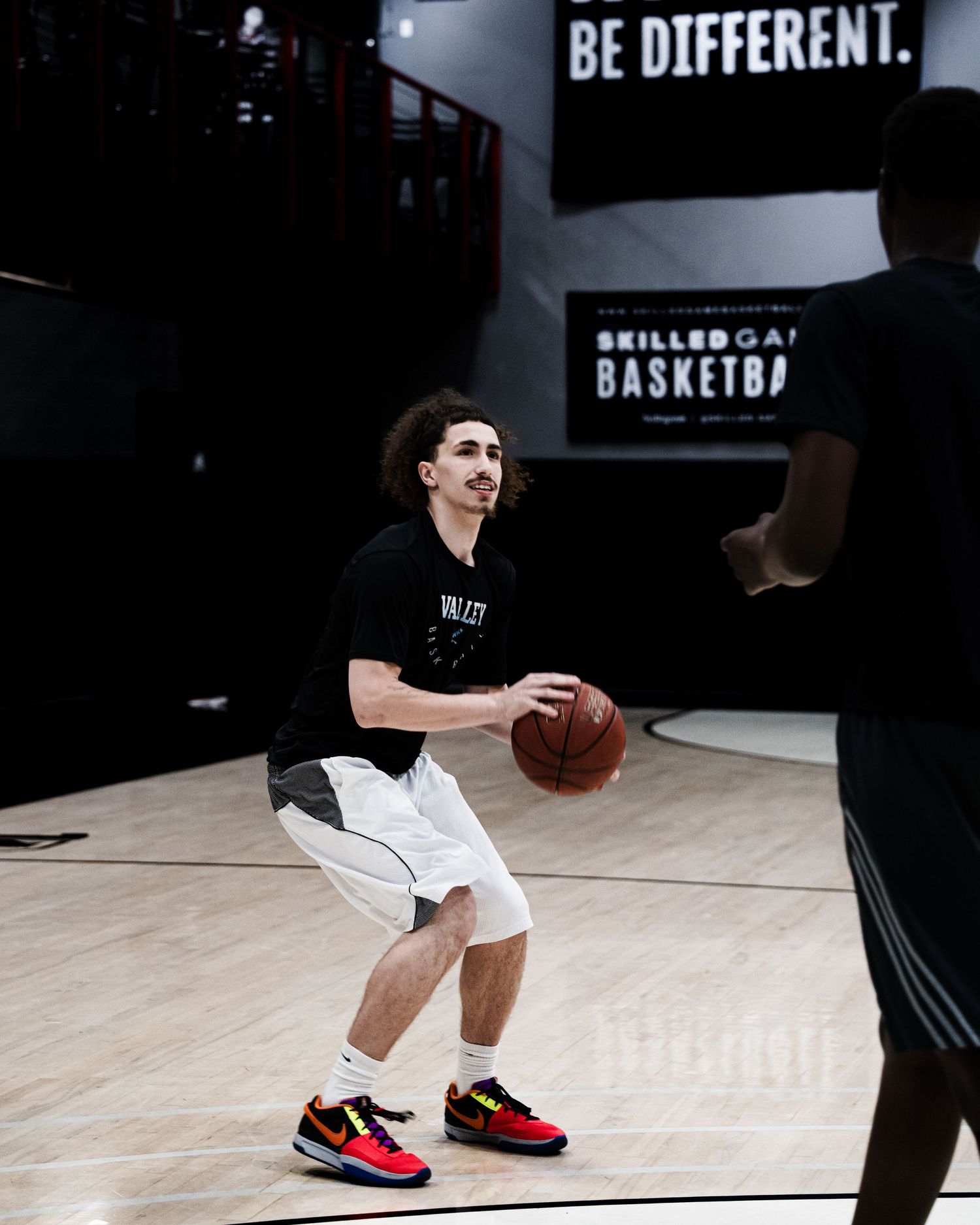 Young man smiling on basketball court holding ball, wearing black tee, white shorts, and colorful sneakers.