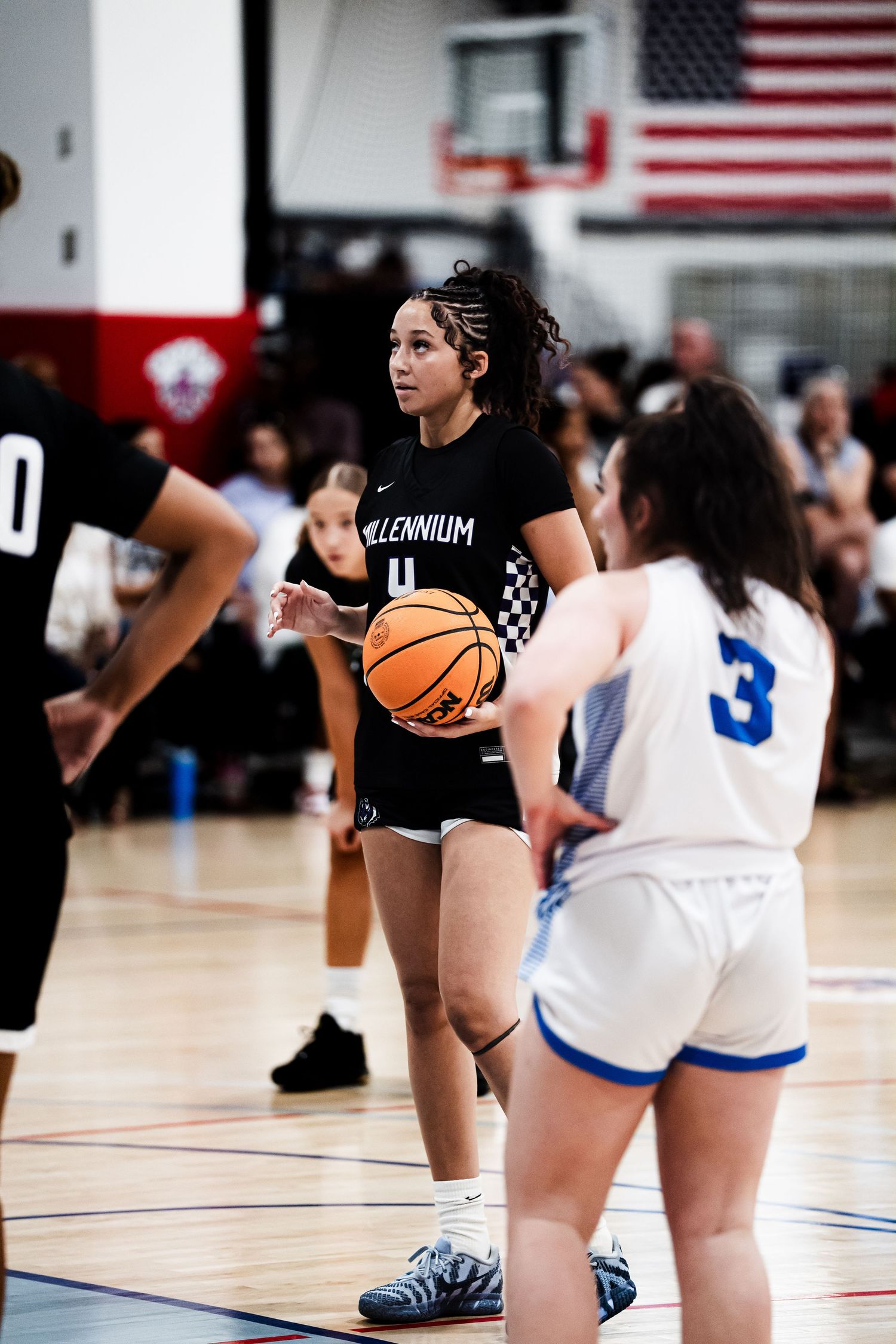 Female basketball player #4 in black Millennium jersey holds ball at free throw line during indoor game.
