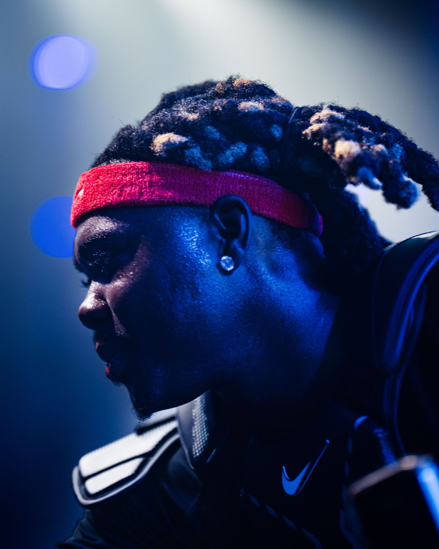 Close-up profile of a young Black man with dreadlocks and red headband performing under blue stage lighting.