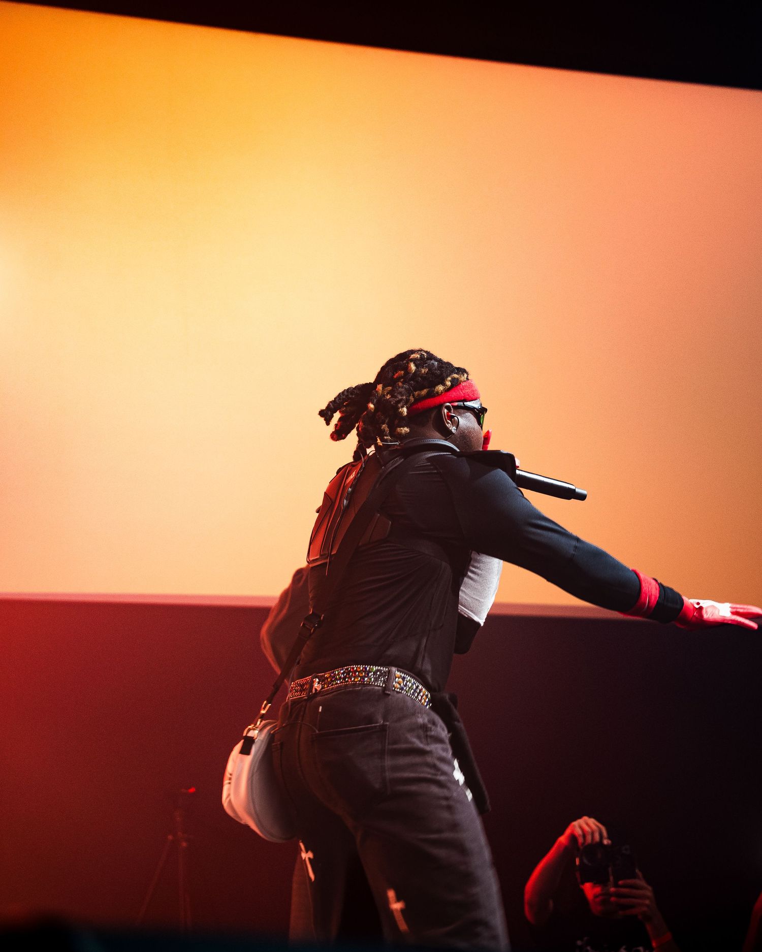 A performer on stage holds a microphone while wearing dreads and a red headband against an orange backdrop.