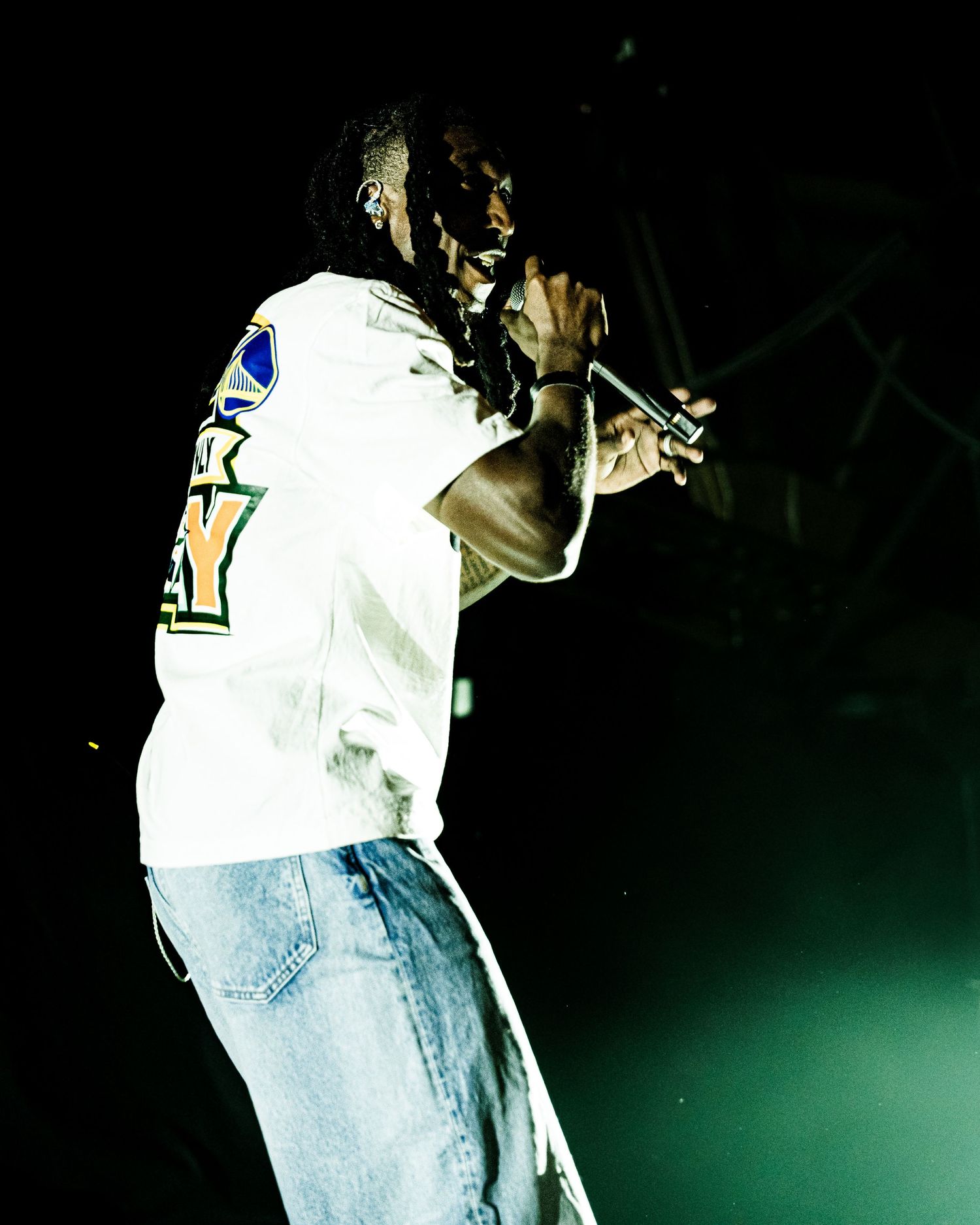 Rapper in white jersey and jeans performs with microphone under green stage spotlight at concert.