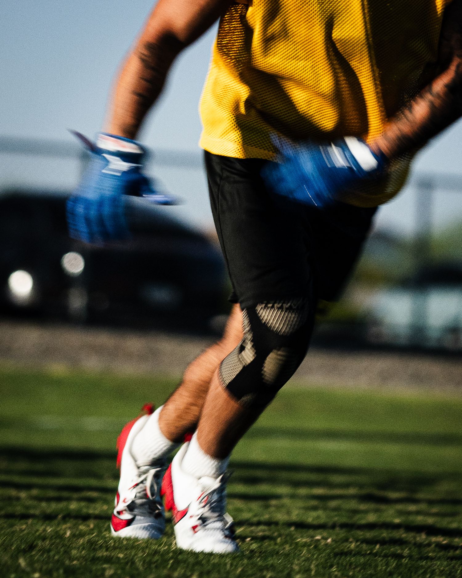 Athlete in yellow jersey and black shorts sprints on grass field wearing blue gloves and red-white cleats.