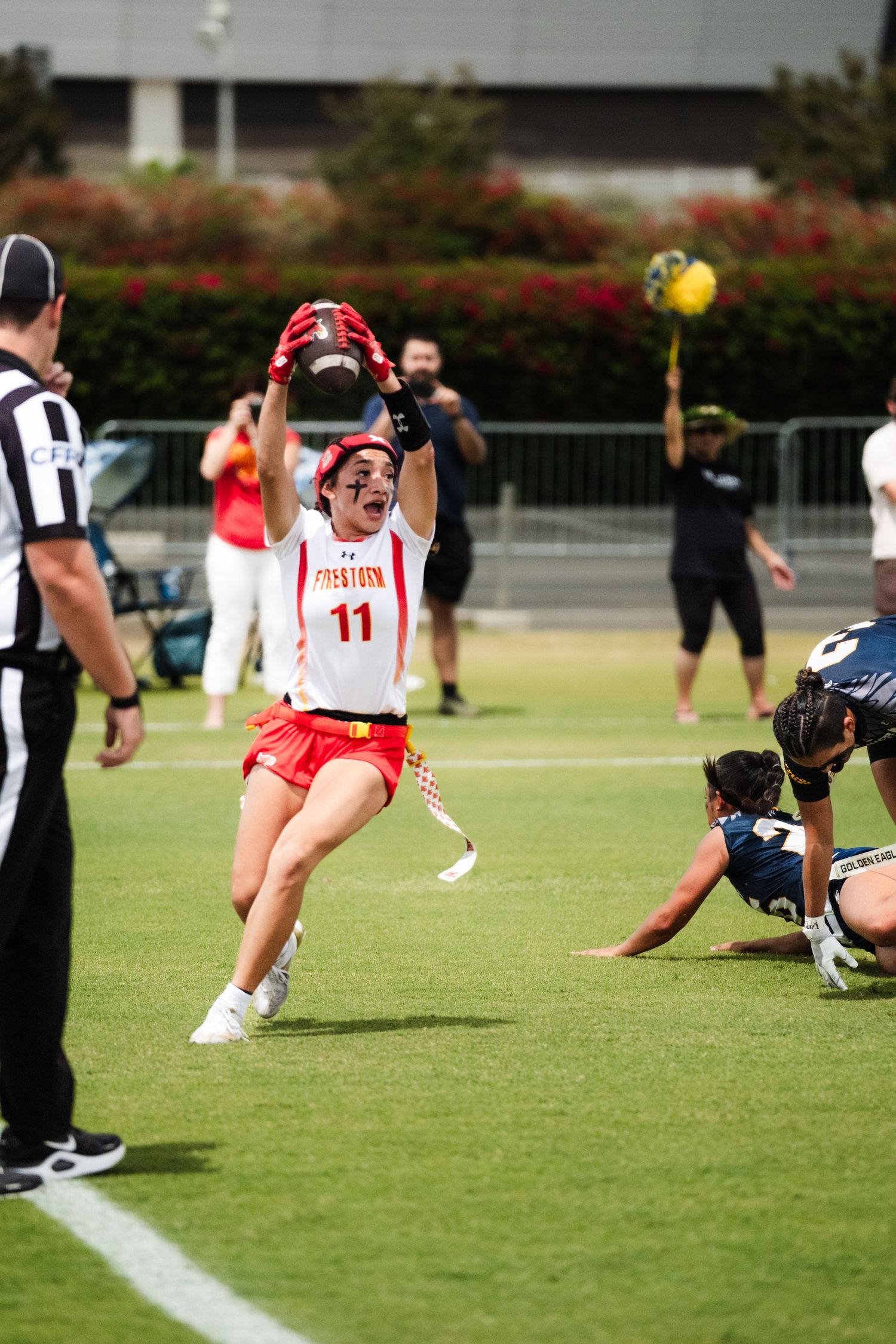 Female lacrosse player #11 in red uniform leaps triumphantly during a college game while referee and opponents watch nearby.