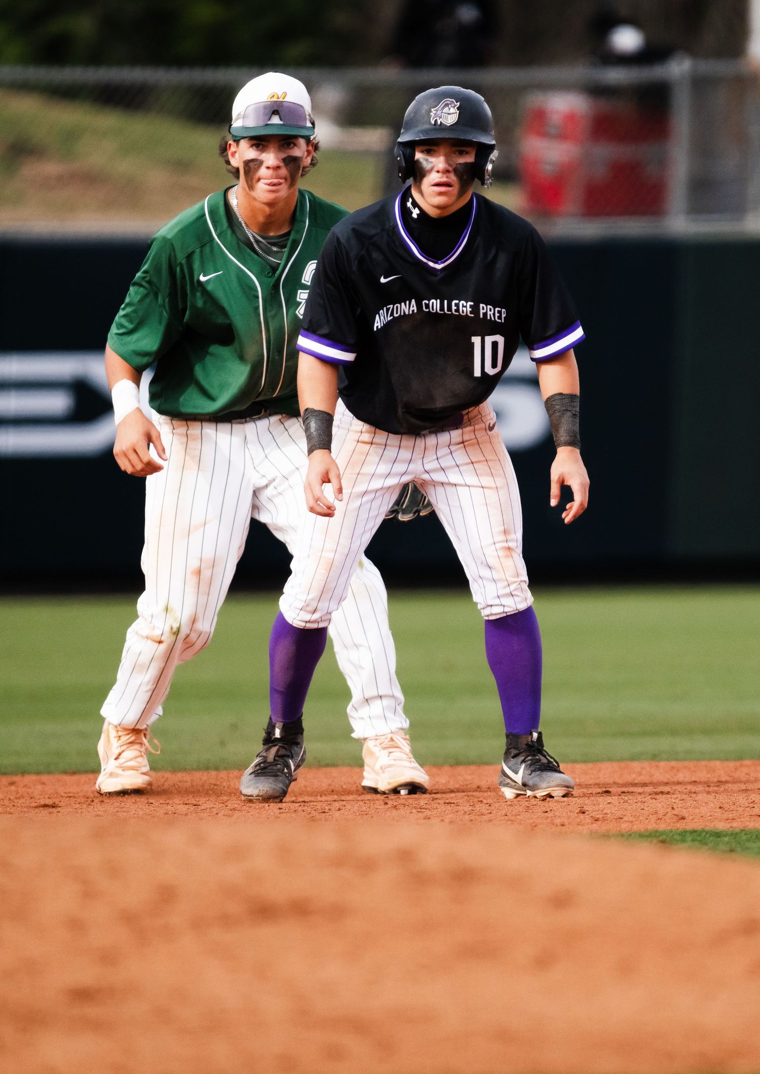 Baseball player #10 in Arizona College Prep uniform leads off base during a college baseball game.