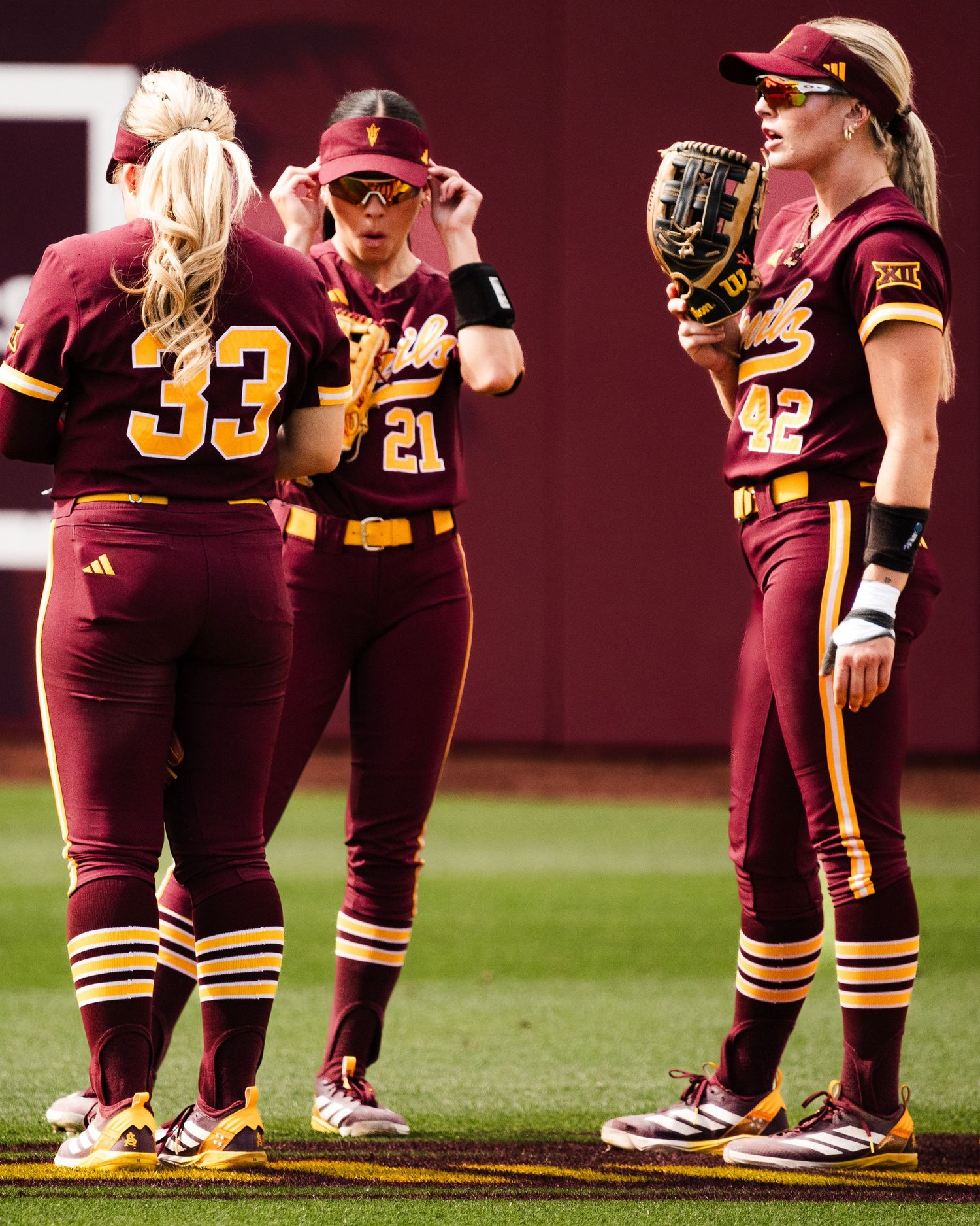 Three Arizona State Sun Devils softball players in maroon uniforms huddle on the field during a Big 12 game.