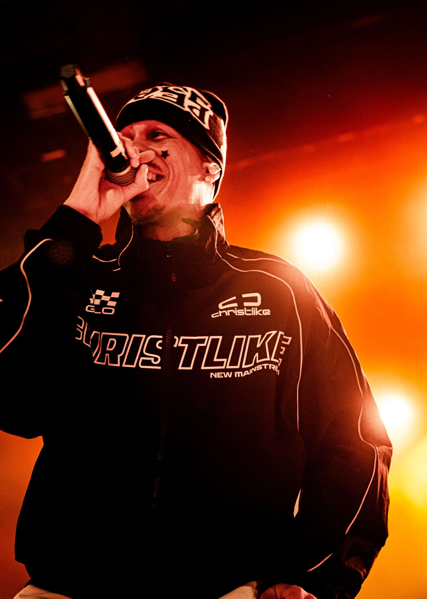 Rapper performing on stage with microphone, wearing black jacket and cap under dramatic orange stage lighting.