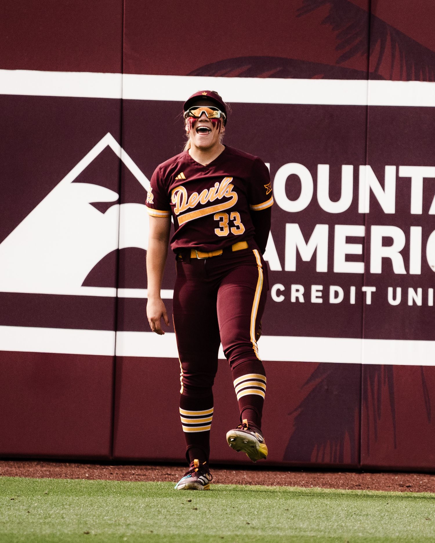 ASU softball player #33 in maroon uniform celebrates on field near Country America Credit Union outfield wall.