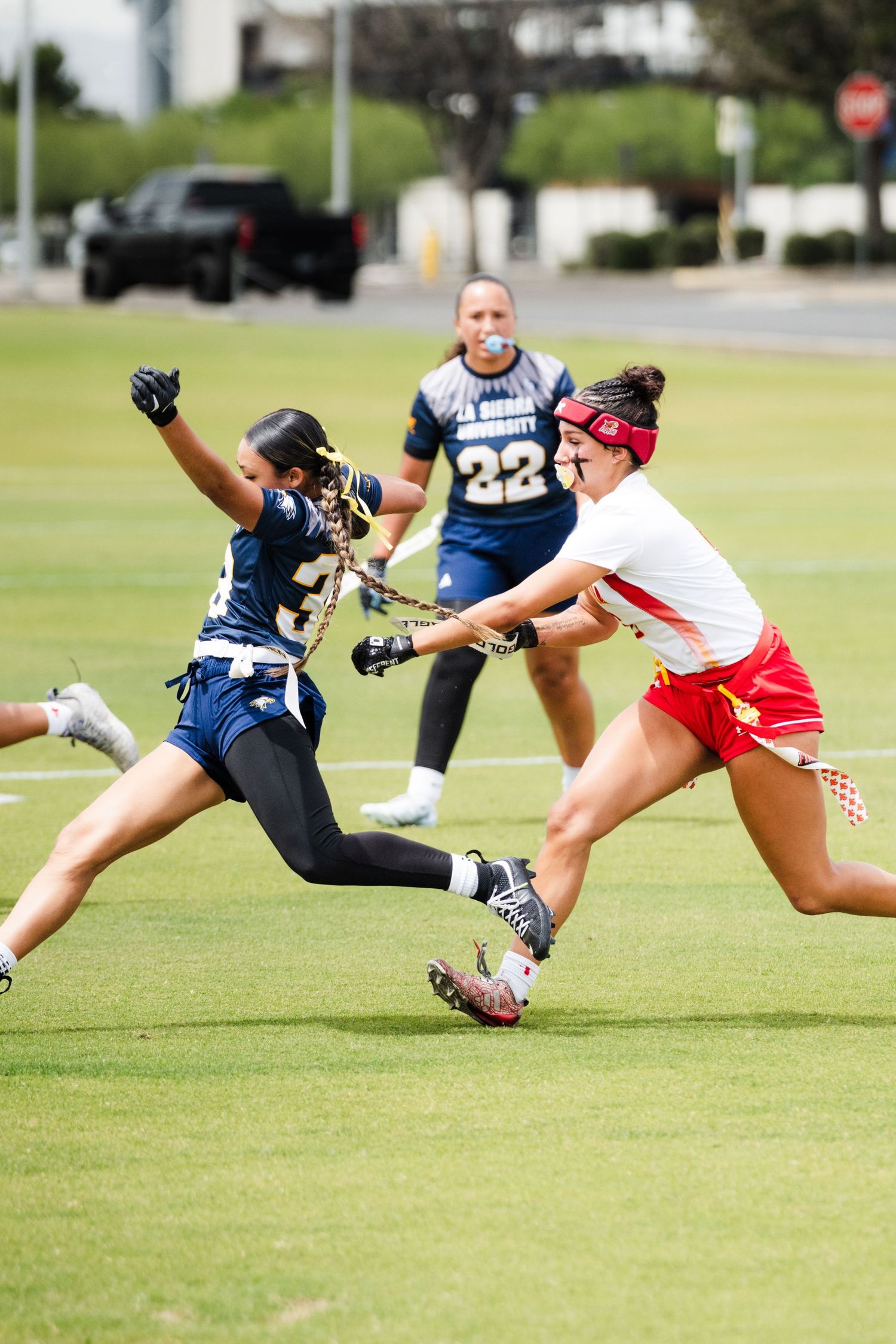 Female lacrosse players compete in an intense game, with one player in red challenging a defender in navy blue uniform.