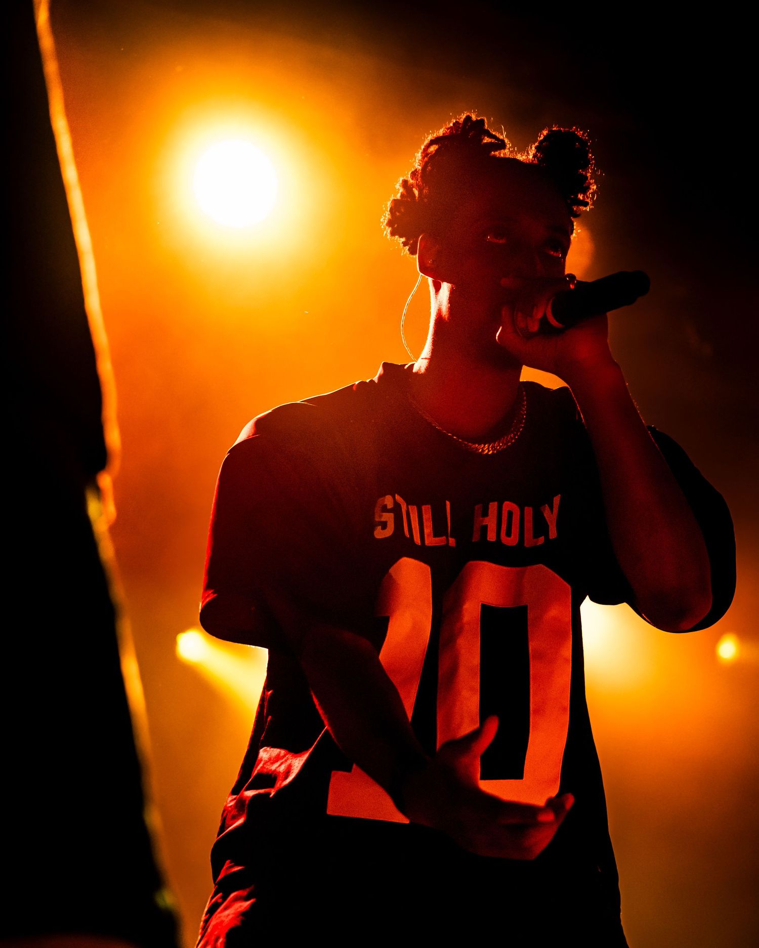 Performer in a Still Holy jersey holds a microphone on a dimly lit stage bathed in warm orange spotlight.
