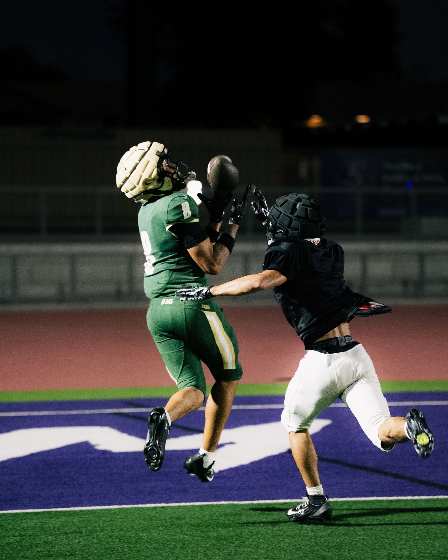 Two football players jump for a pass under stadium lights during a night game, one in green and one in black.