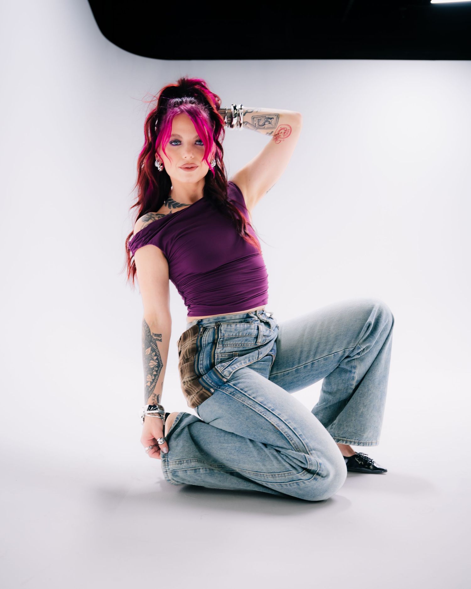 Woman with pink hair and tattoos poses in purple crop top and wide-leg jeans on white studio backdrop.