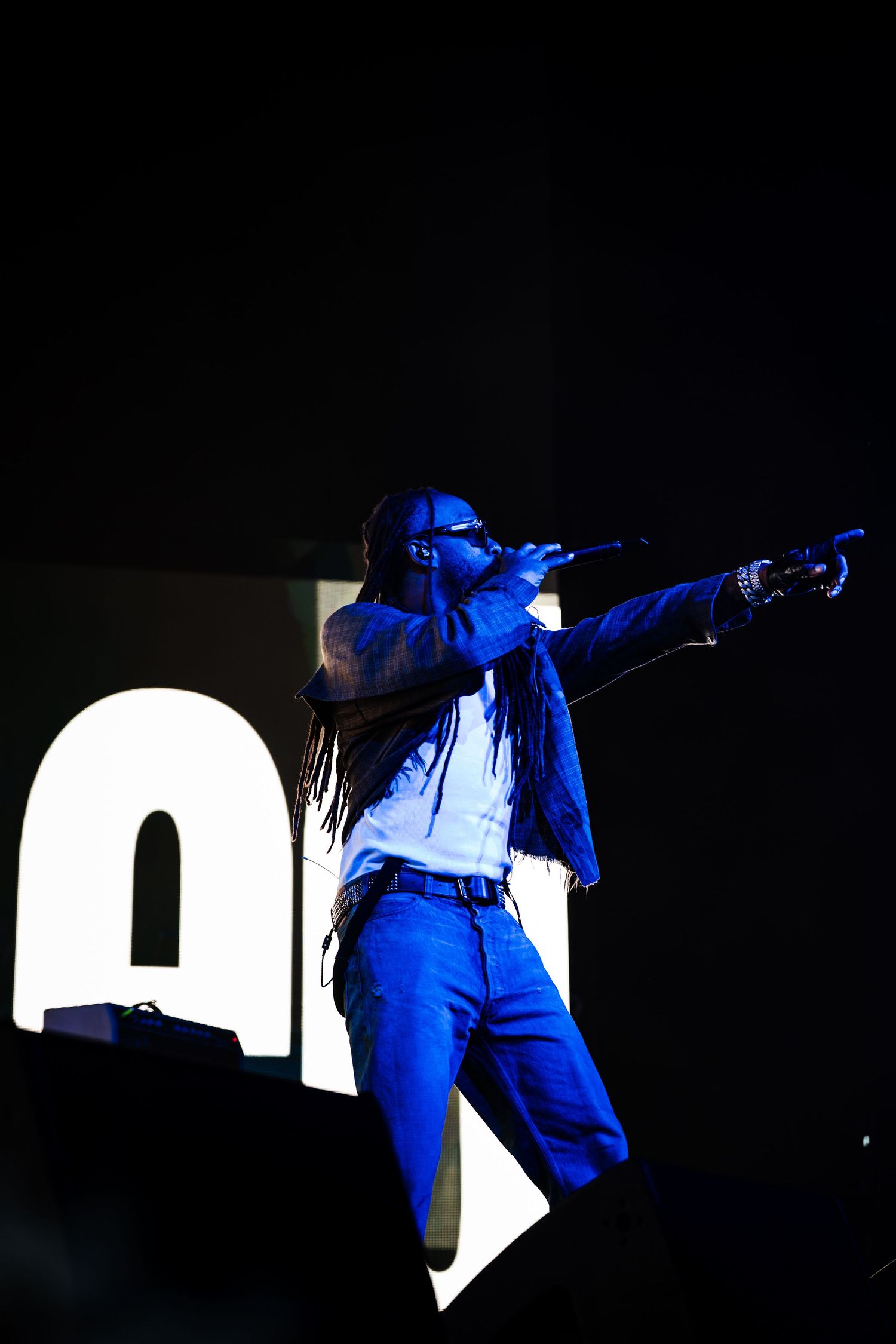 Performer with long dreadlocks and sunglasses sings into a microphone and points outward on a dark stage bathed in blue light.