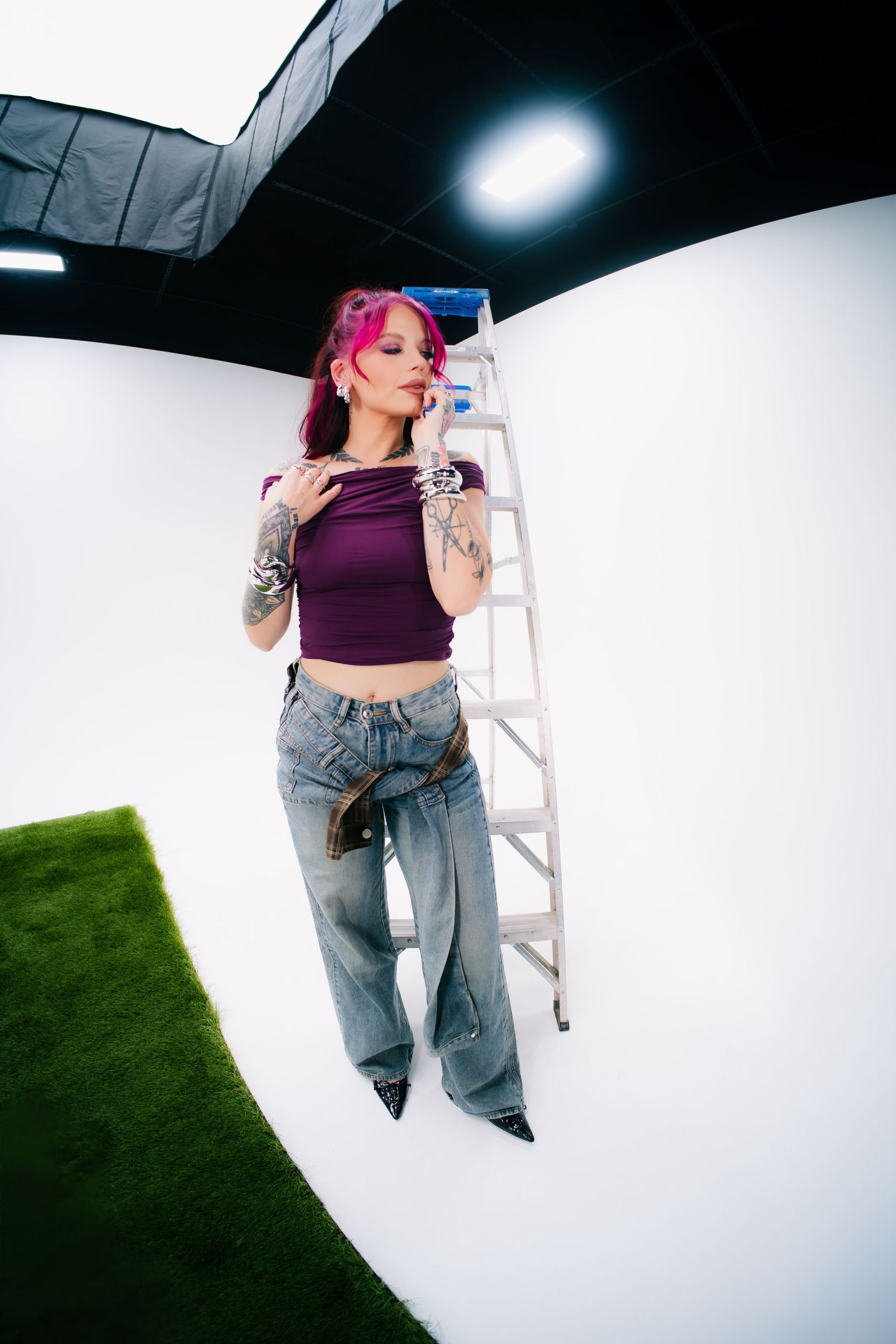 Tattooed woman with pink hair in purple crop top and wide-leg jeans poses by a ladder in a photo studio.