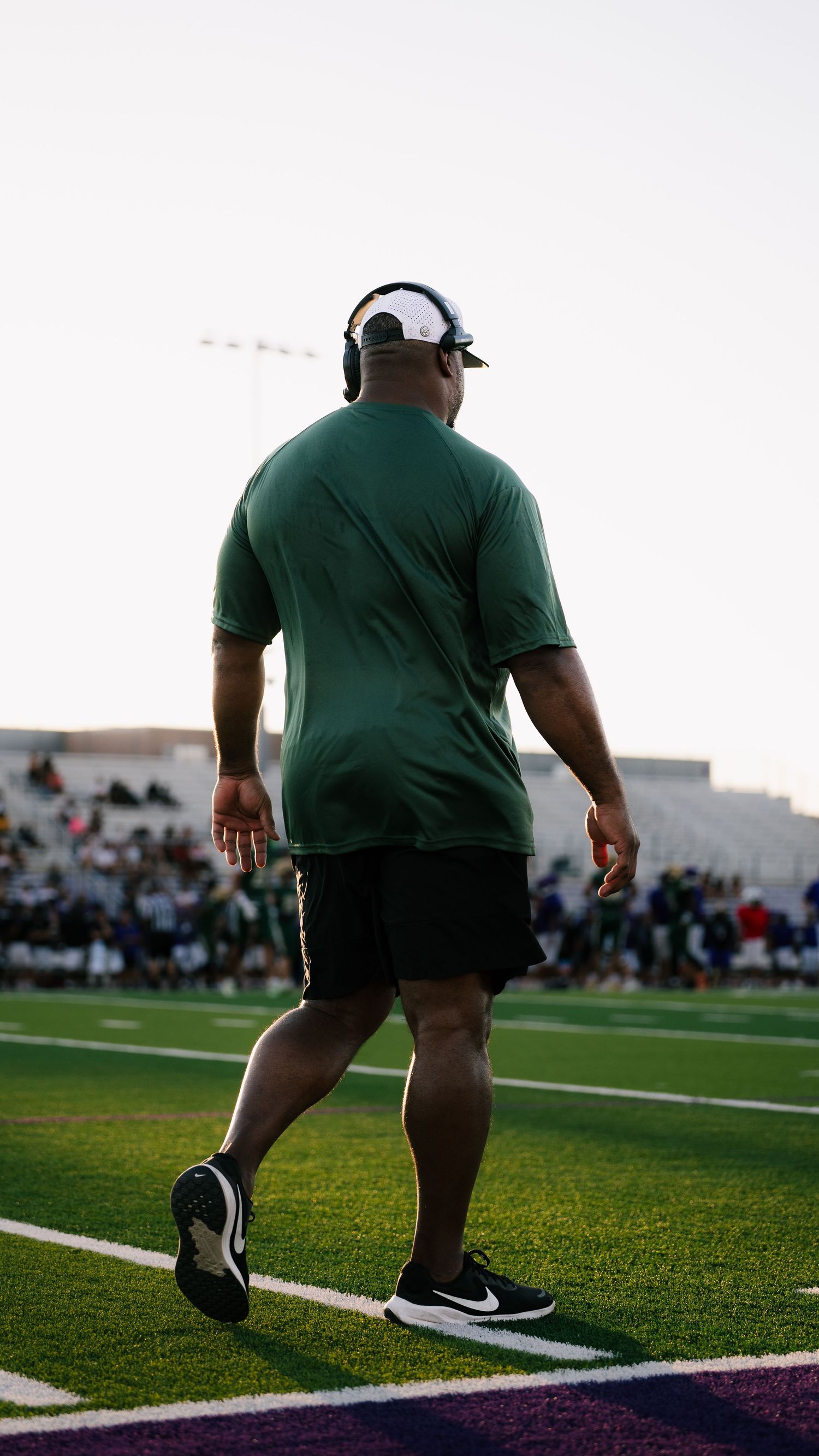 A football coach in green shirt and black shorts walks the sideline during a high school football game.