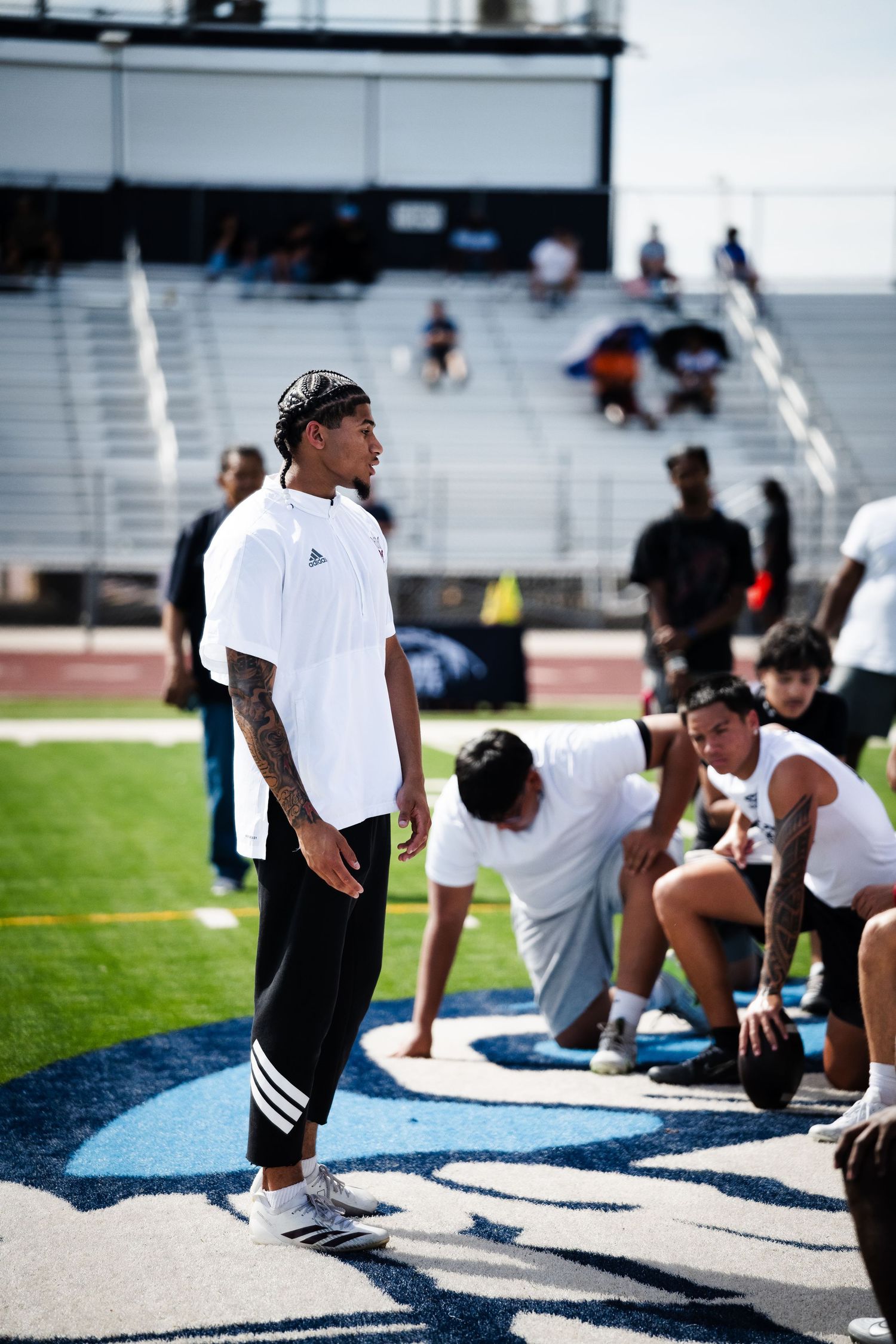Tattooed athlete in Adidas gear stands on a football field logo during a training camp event.