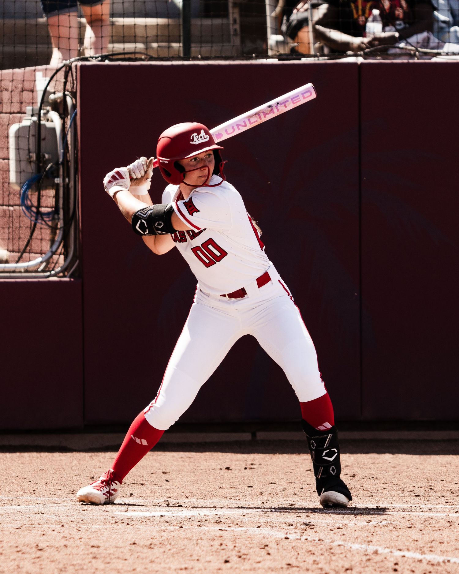 College softball player in white uniform assumes batting stance holding pink bat on dirt field.