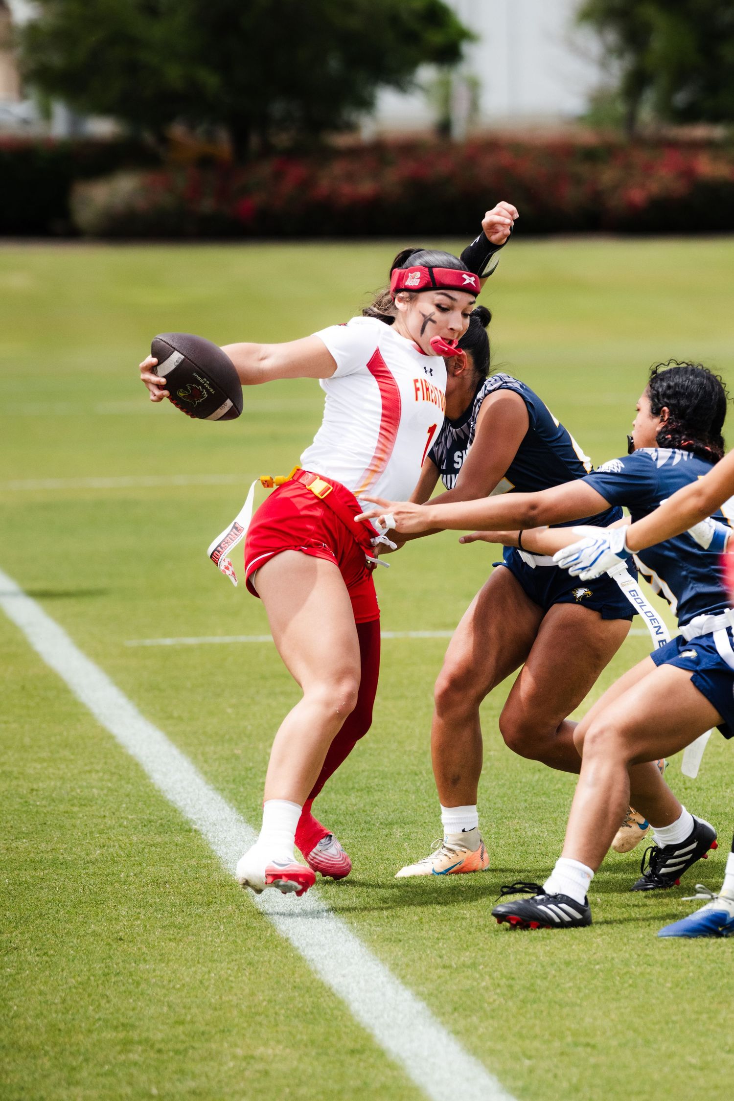 Female flag football player in white and red uniform prepares to throw ball while defenders attempt to pull her flag.