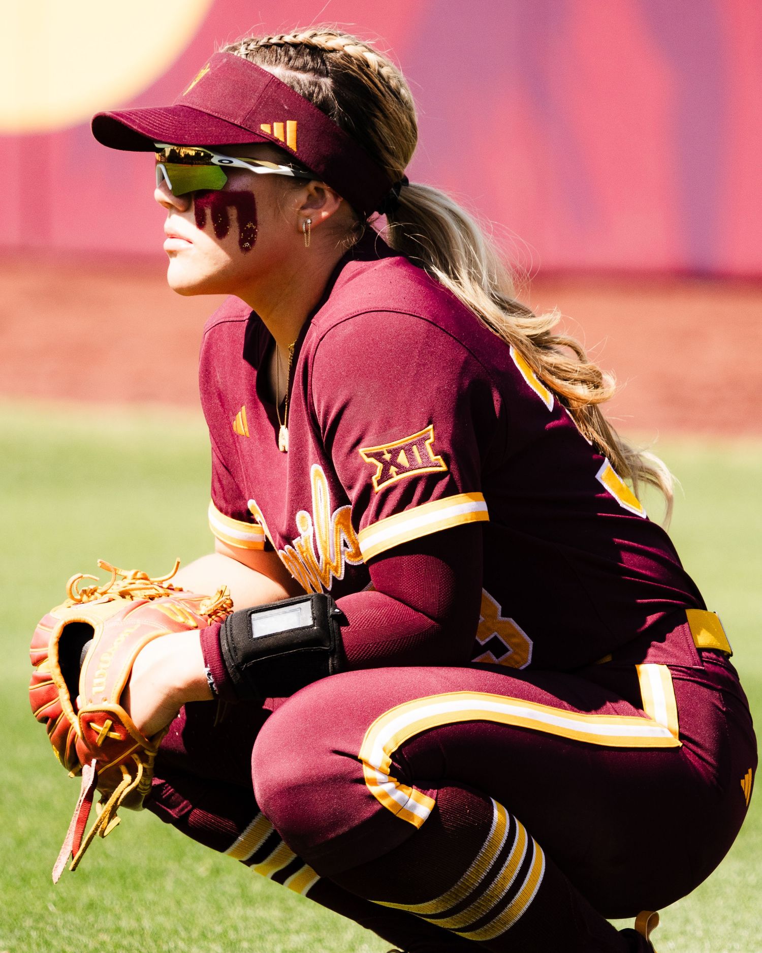 Female softball player in maroon Big 12 uniform crouching with glove, wearing sunglasses and cap on sunny field.