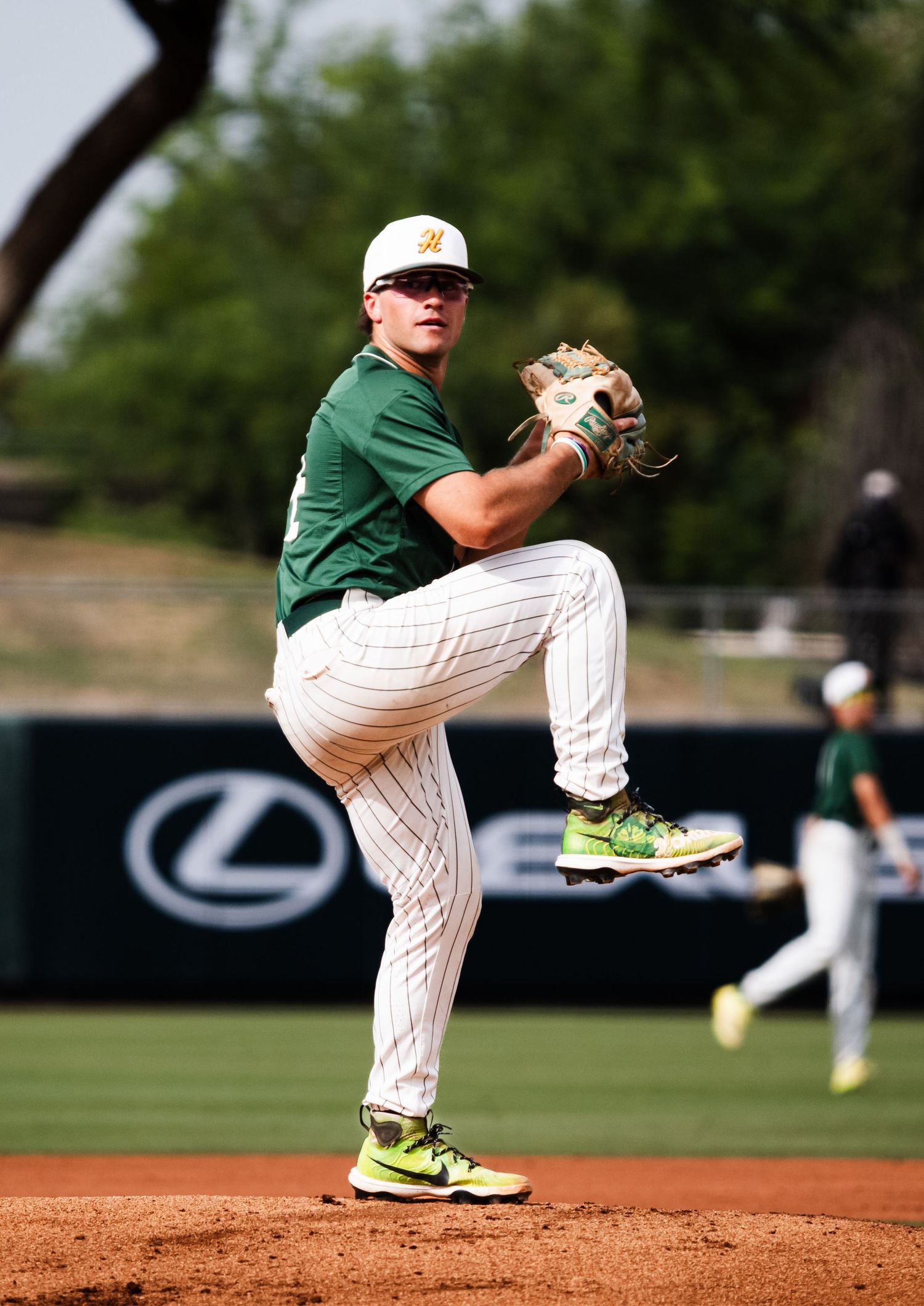 Baseball pitcher in green uniform mid-windup on mound, wearing yellow cleats, with Lexus advertisement in background.