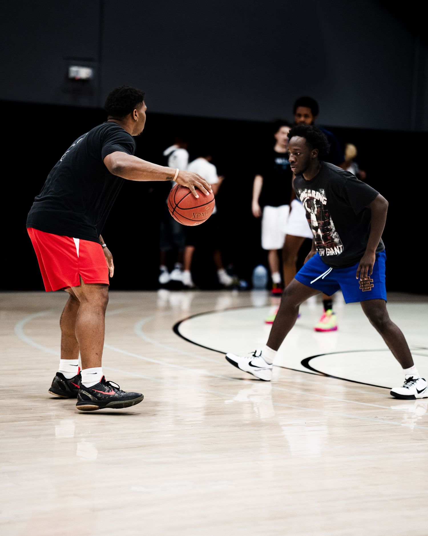 Two basketball players practice defensive drills on a bright indoor hardwood court.