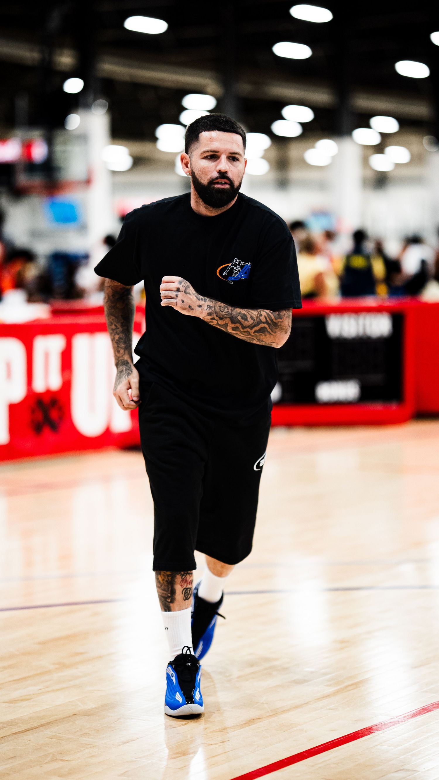 Tattooed man in black shirt and shorts jogging on an indoor basketball court wearing blue and white sneakers.
