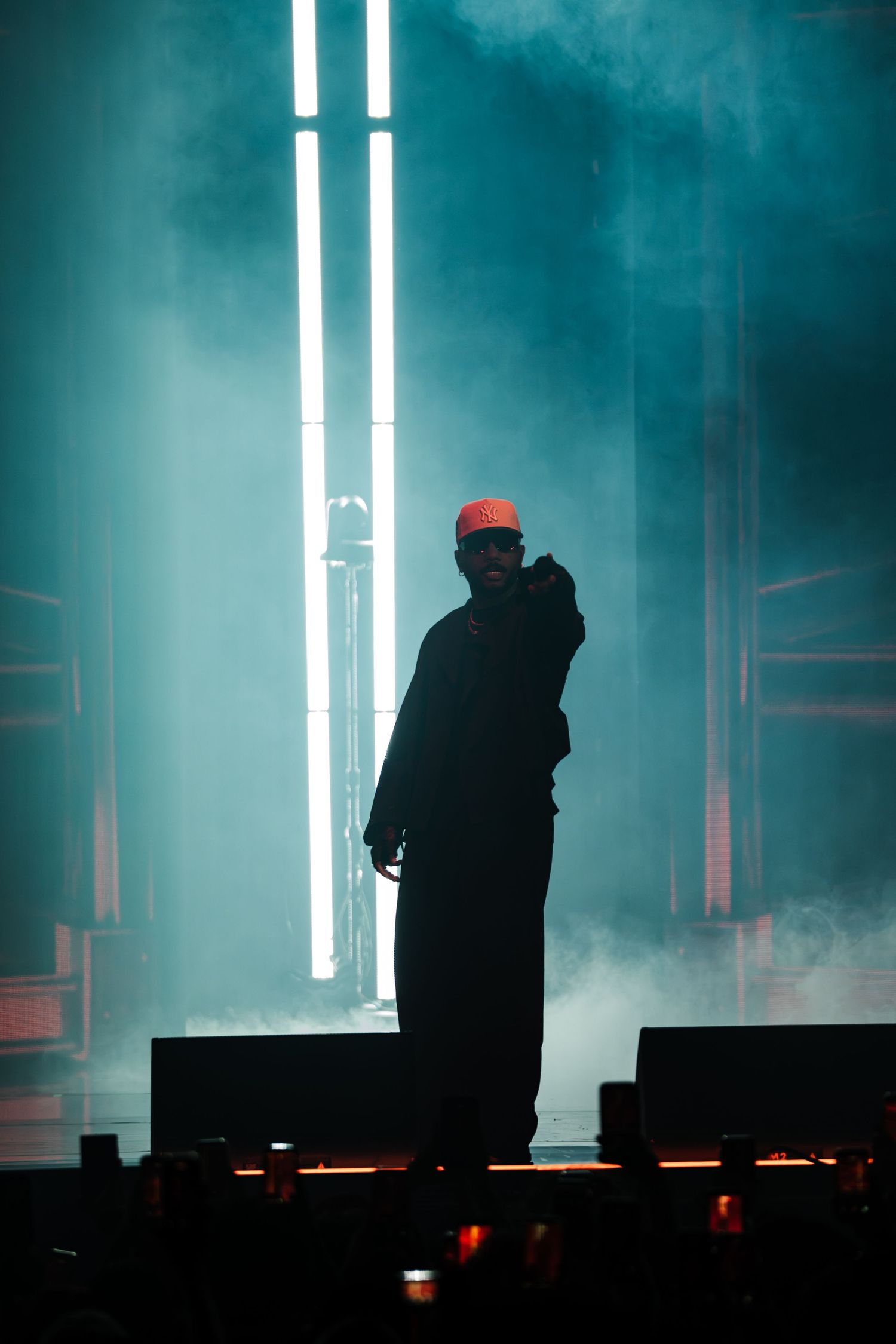 Silhouetted performer in an orange cap and dark clothing points toward the crowd on a smoky stage lit by tall vertical white lights.