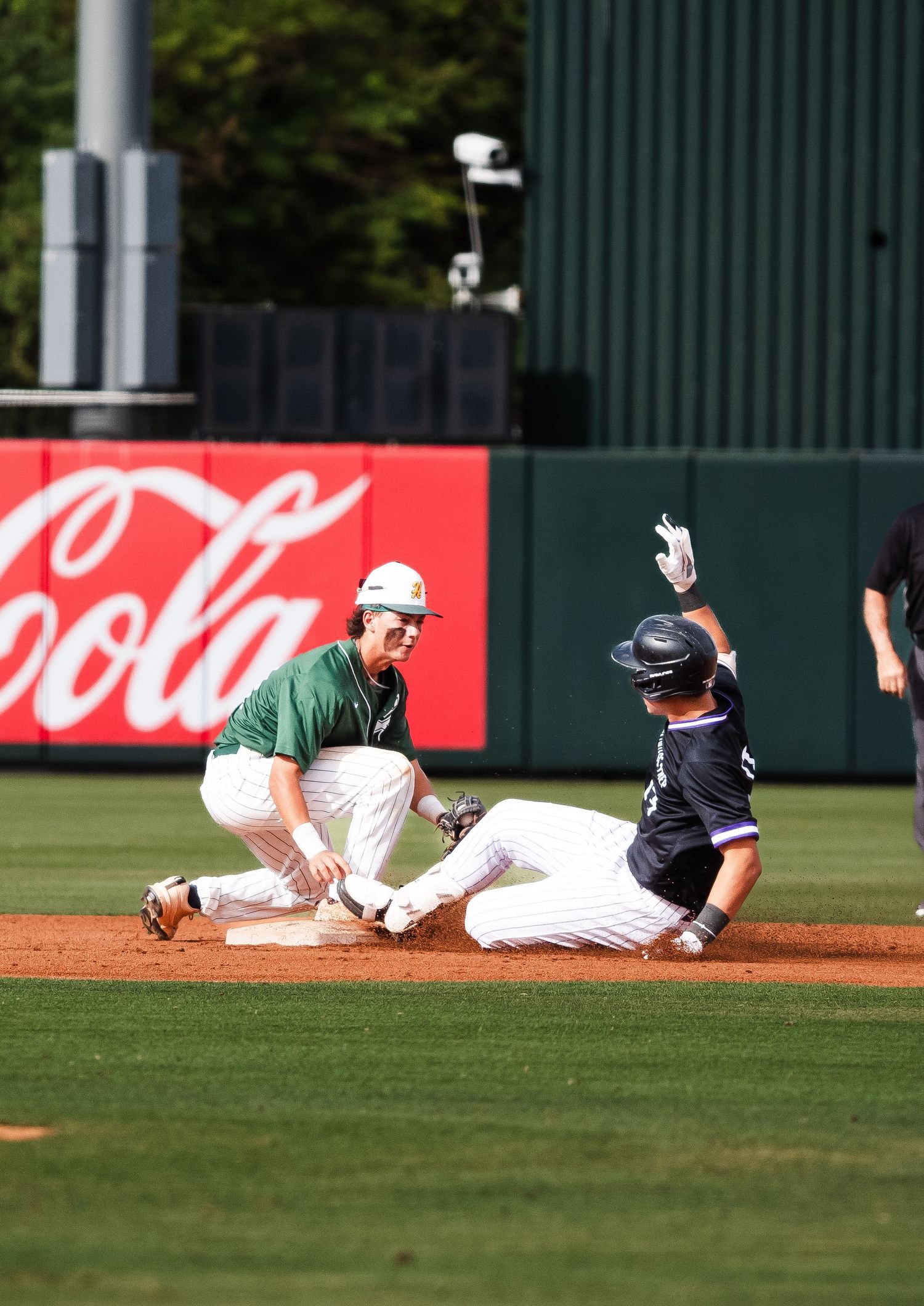 Baseball player slides into second base as green-uniformed infielder applies tag during game near Coca-Cola wall.