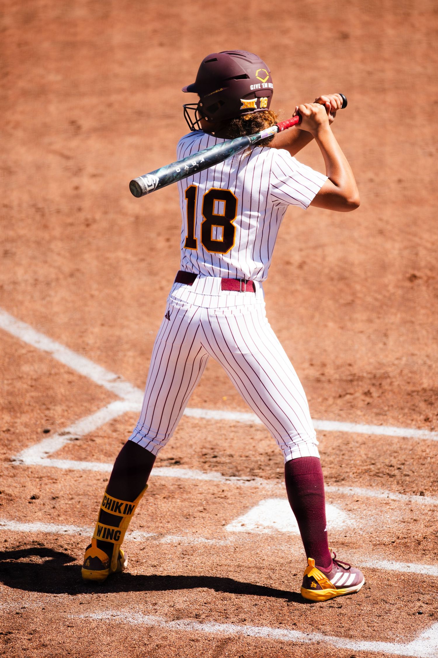 Female softball player number 18 in maroon pinstripe uniform stands ready at bat during a college game.