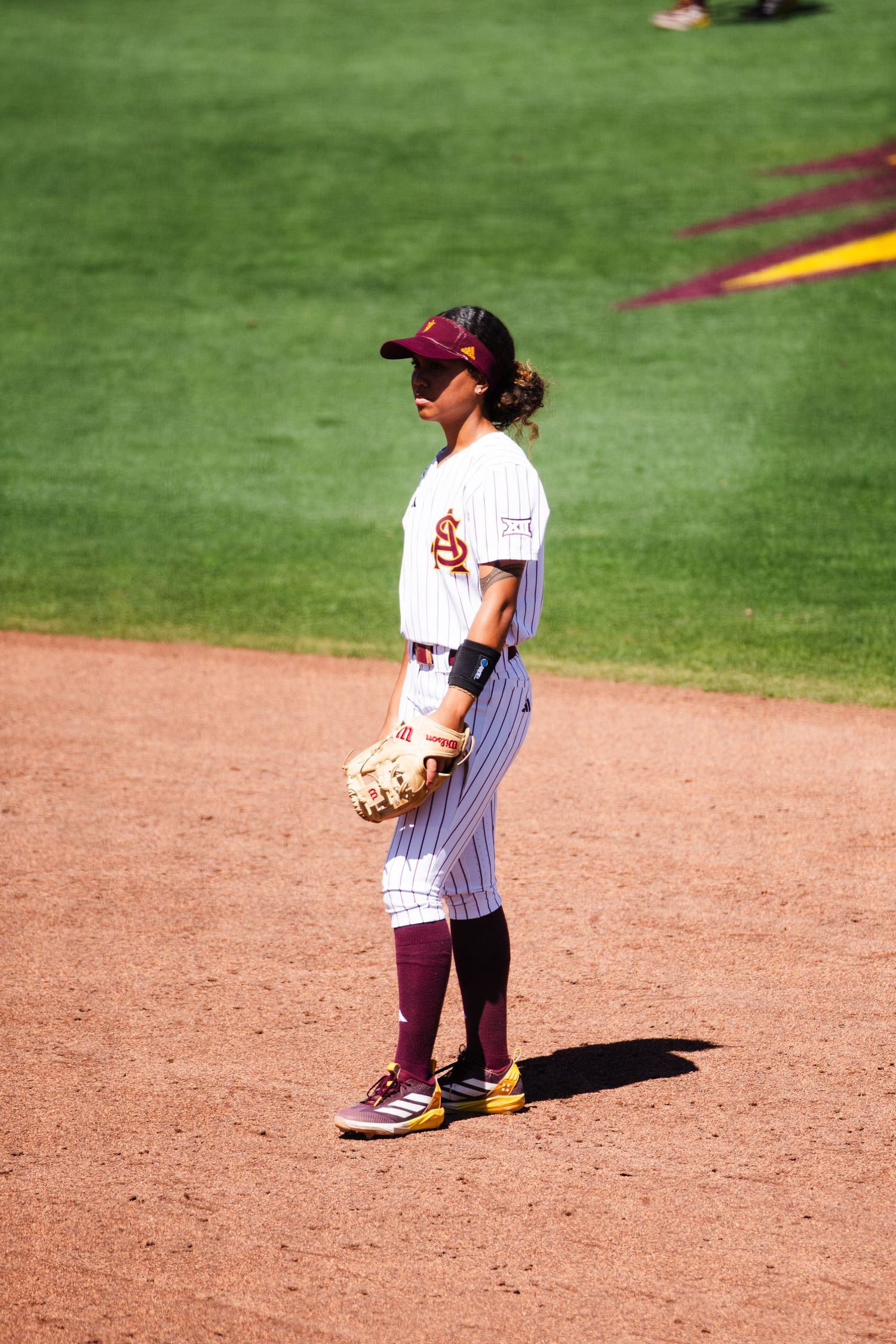Arizona State Sun Devils softball player stands ready on the field in white pinstripe uniform and maroon cap.
