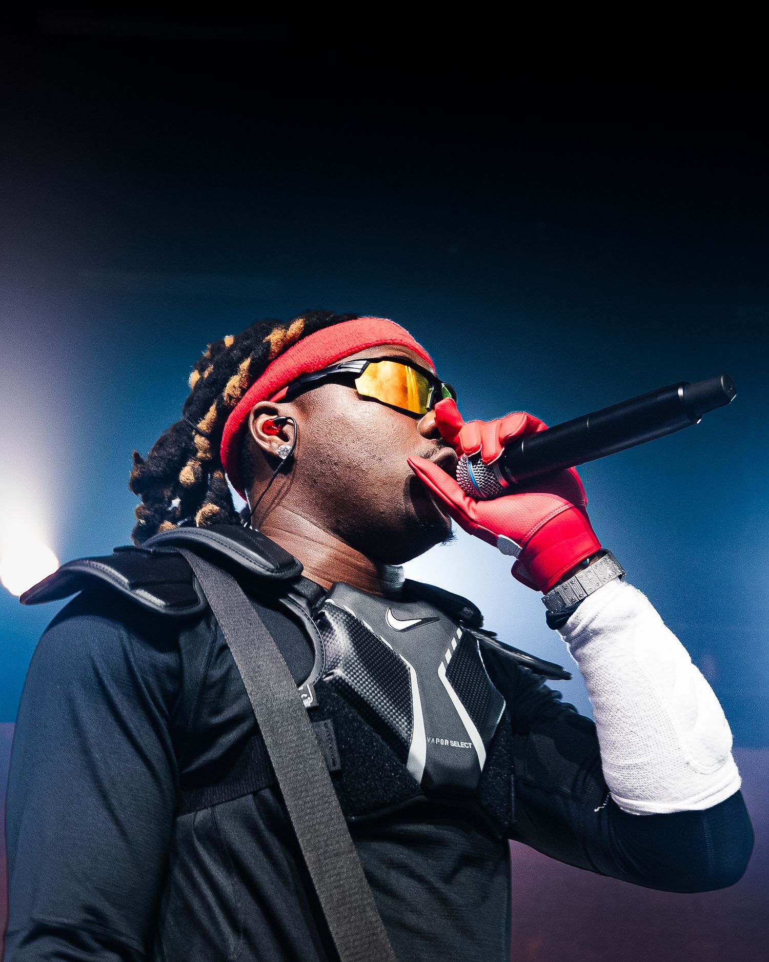 Rapper performing live on stage wearing red headband, Nike gloves, and yellow tinted sunglasses while holding a microphone.