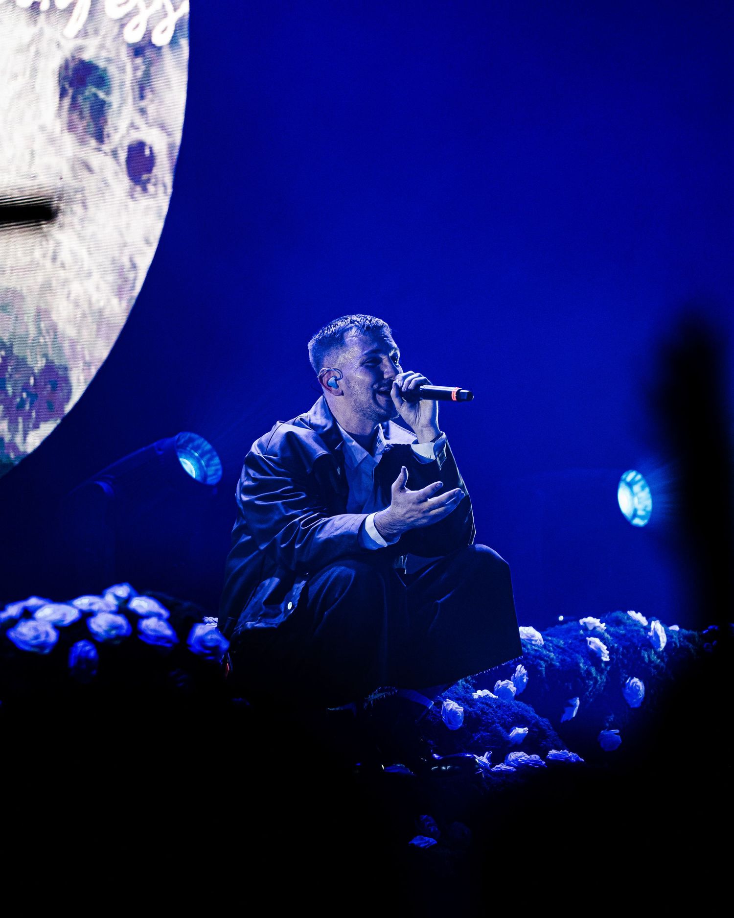 Singer performing on stage bathed in blue light, kneeling among flowers with a large moon projection backdrop.