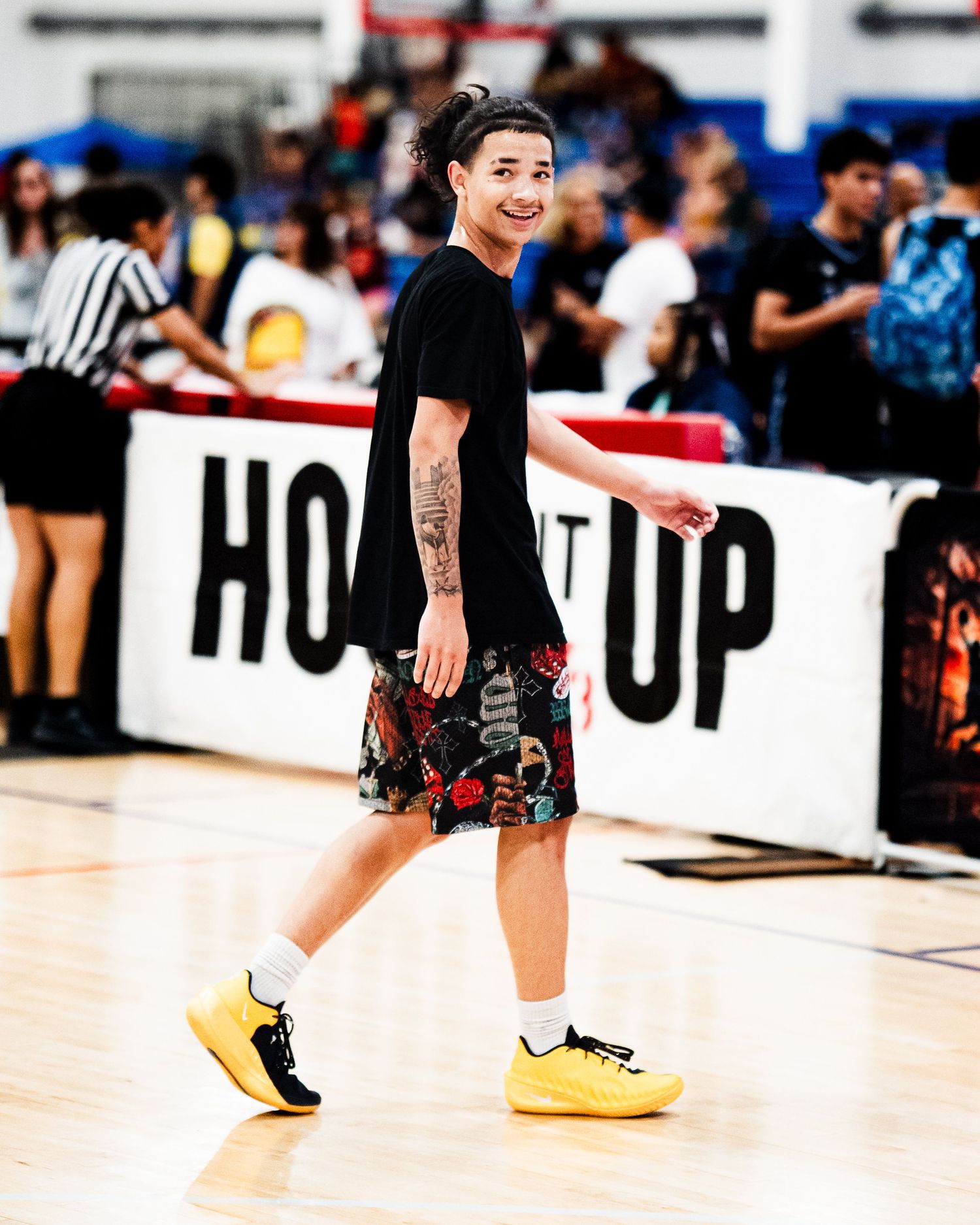 A smiling young person walks on a basketball court wearing a black tee, floral shorts, and yellow sneakers during an event.