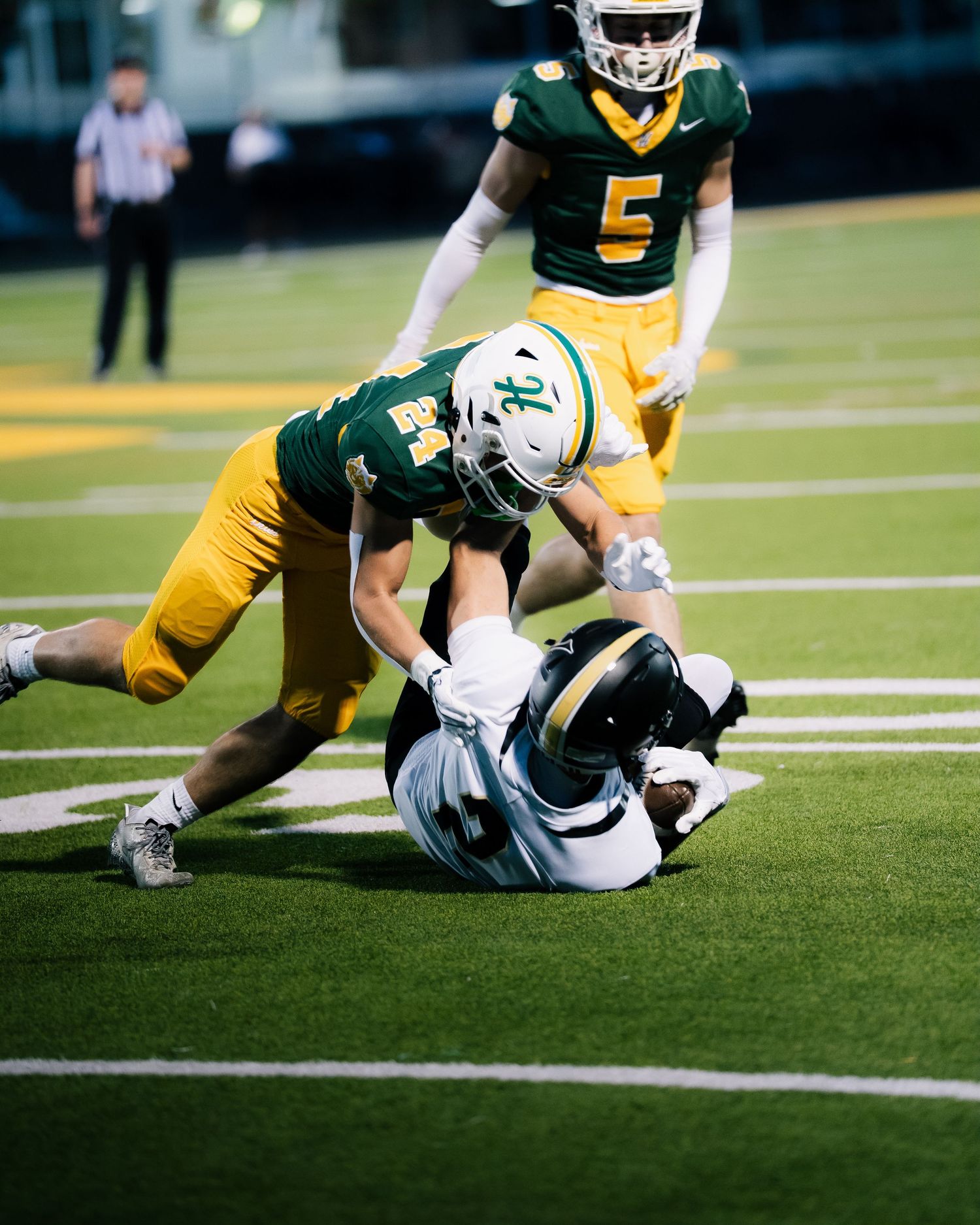 Football player tackled on field by green and gold uniformed defender during a night game.