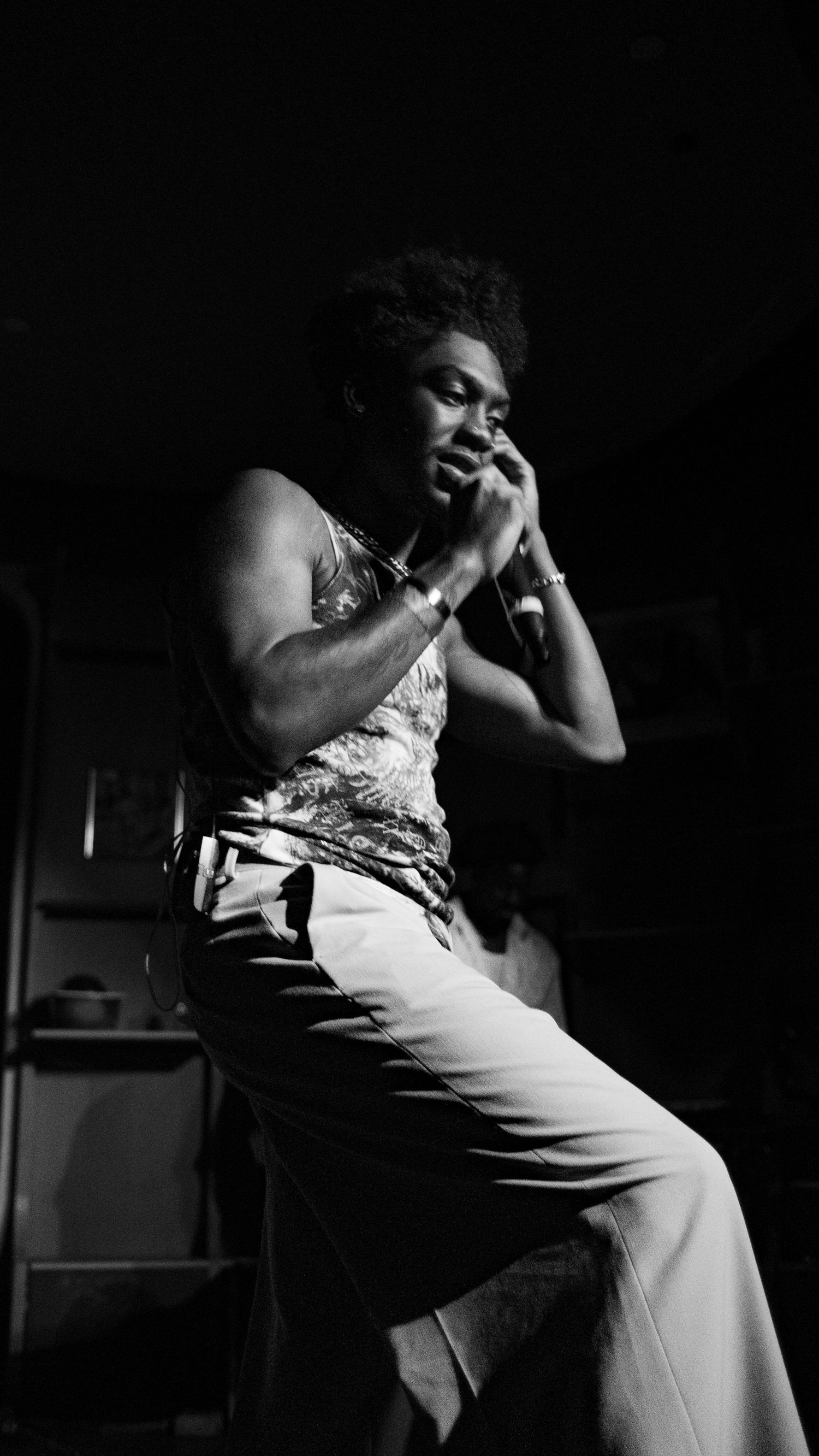 Black and white photo of a young man sitting on stage, holding a microphone close to his mouth during a live performance.